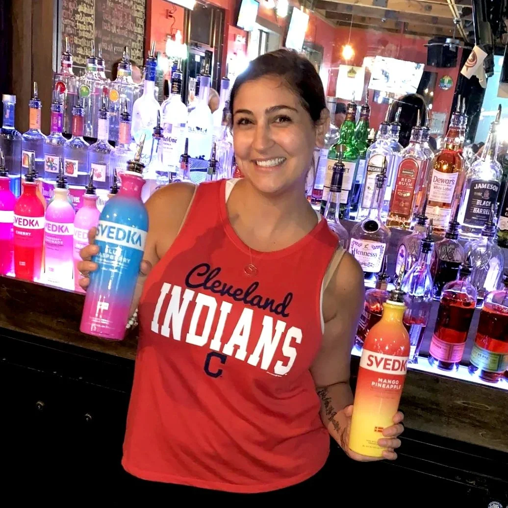 Woman in a Cleveland Indians tank top smiling and holding colorful bottles of Svedka flavored vodka in a bar with liquor bottles in the background.