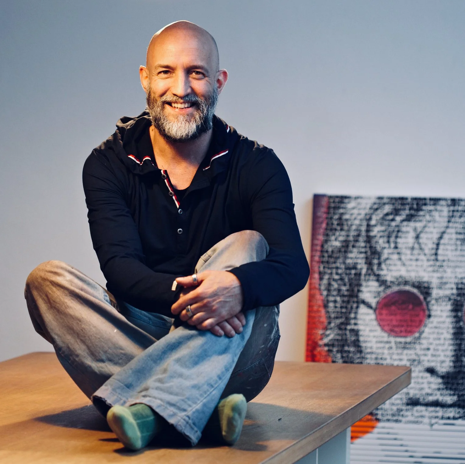Ein Mann mit Bart sitzt auf einem Holztisch, trägt schwarze Kleidung und beige Hosen, mit einem Lächeln im Gesicht vor einer grauen Wand. Hinter ihm steht eine abstrakte Kunstinstallation mit roten, schwarzen und weißen Mustern.