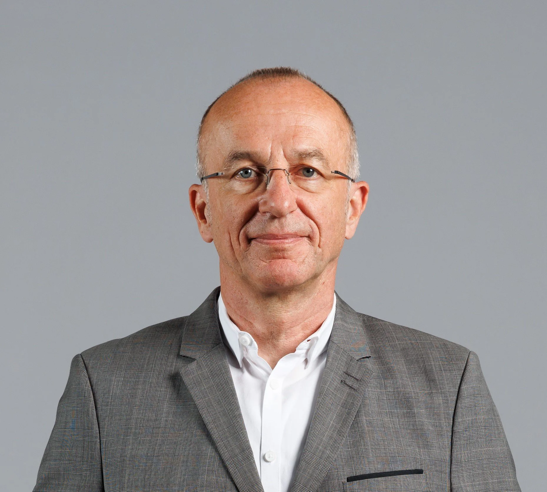 Ein Mann mit Brille trägt einen grauen Anzug und ein weißes Hemd, vor einem grauen Hintergrund.