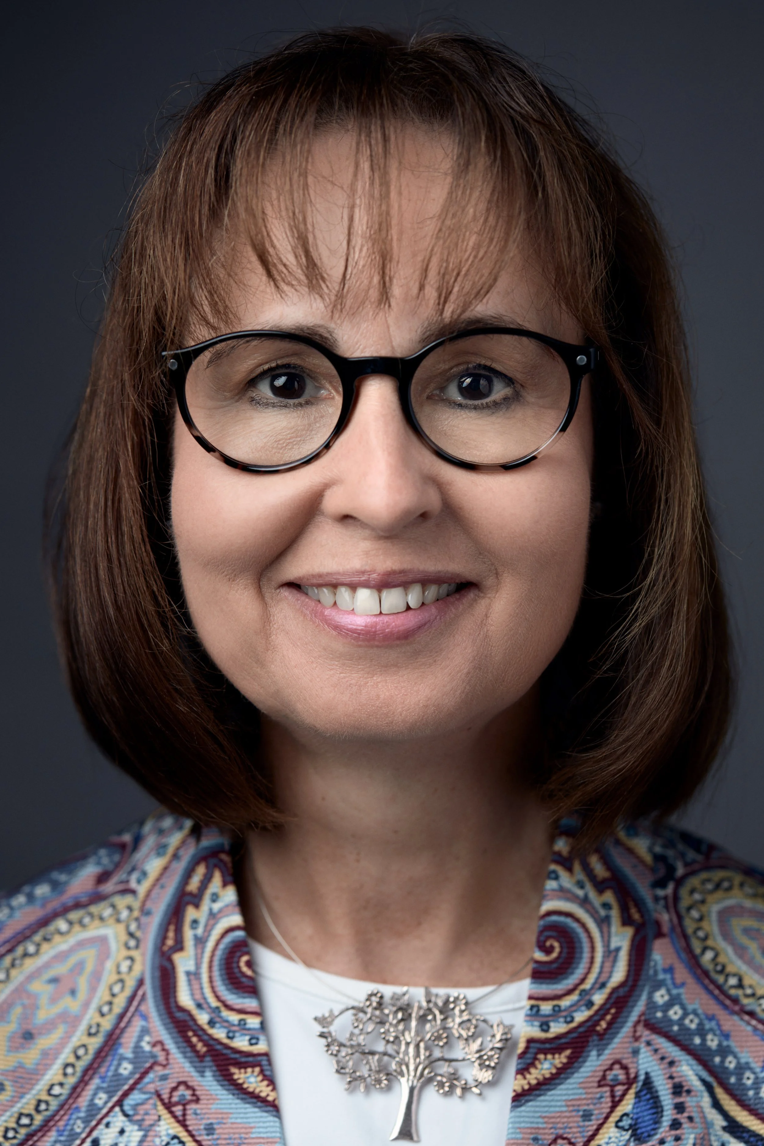 Porträt einer lächelnden Frau mit Brille, braunen Haaren, weißem Top und einer silbernen Baum-Halskette, vor dunklem Hintergrund.