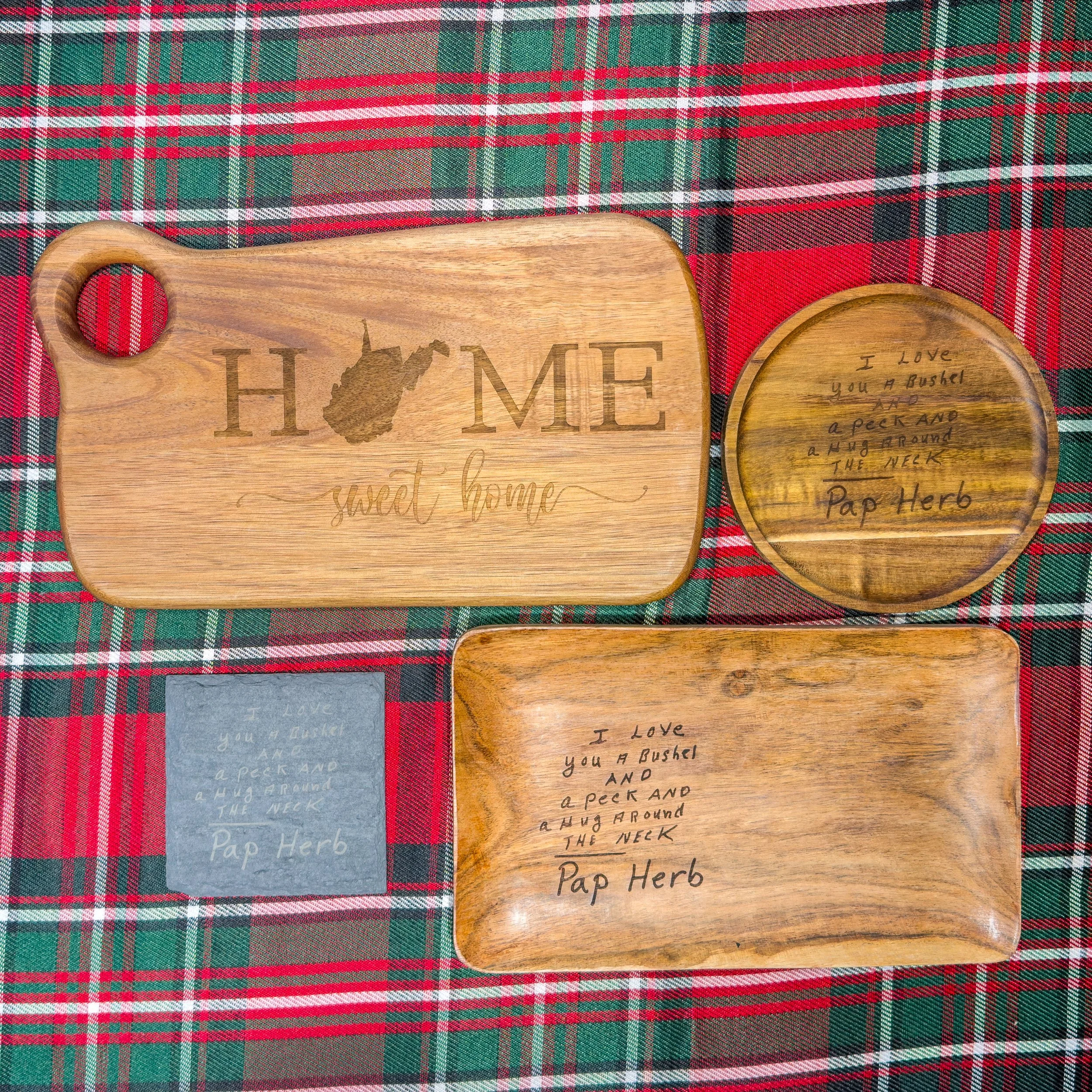 Set of three wooden items placed on red, green, and black plaid fabric: a rectangular cutting board with "HOME sweet home" engraved, a round plate with handwritten message, and a square slate with handwritten message.