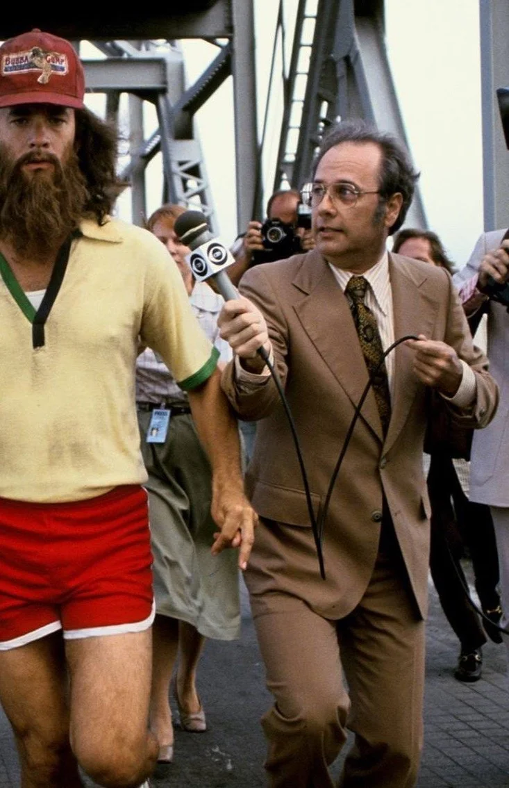 A man with long hair and a beard, wearing a yellow shirt and red shorts, walks on a bridge. He is being followed by journalists, including one holding a CBS microphone, along with a camera crew.