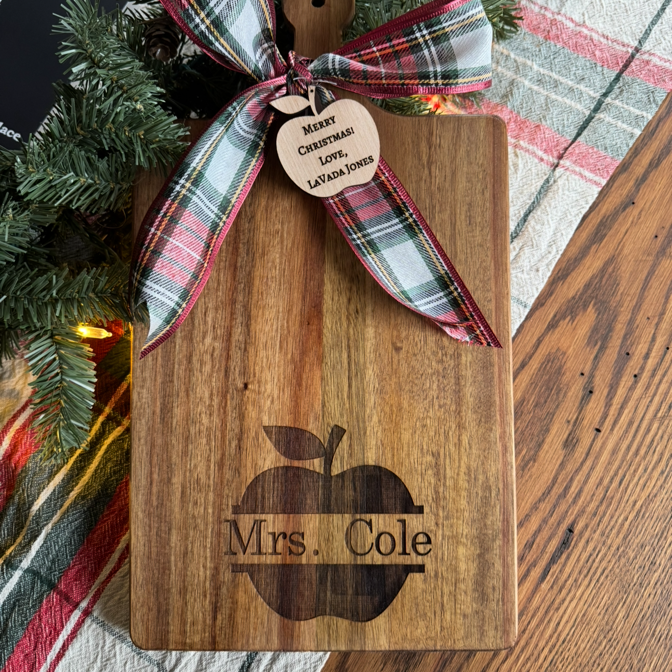 A wooden cutting board with an engraved apple and the text "Mrs. Cole". It is decorated with a plaid red, green, and white ribbon bow, a wooden tag with a message for Christmas, and some pine tree branches with lights nearby.