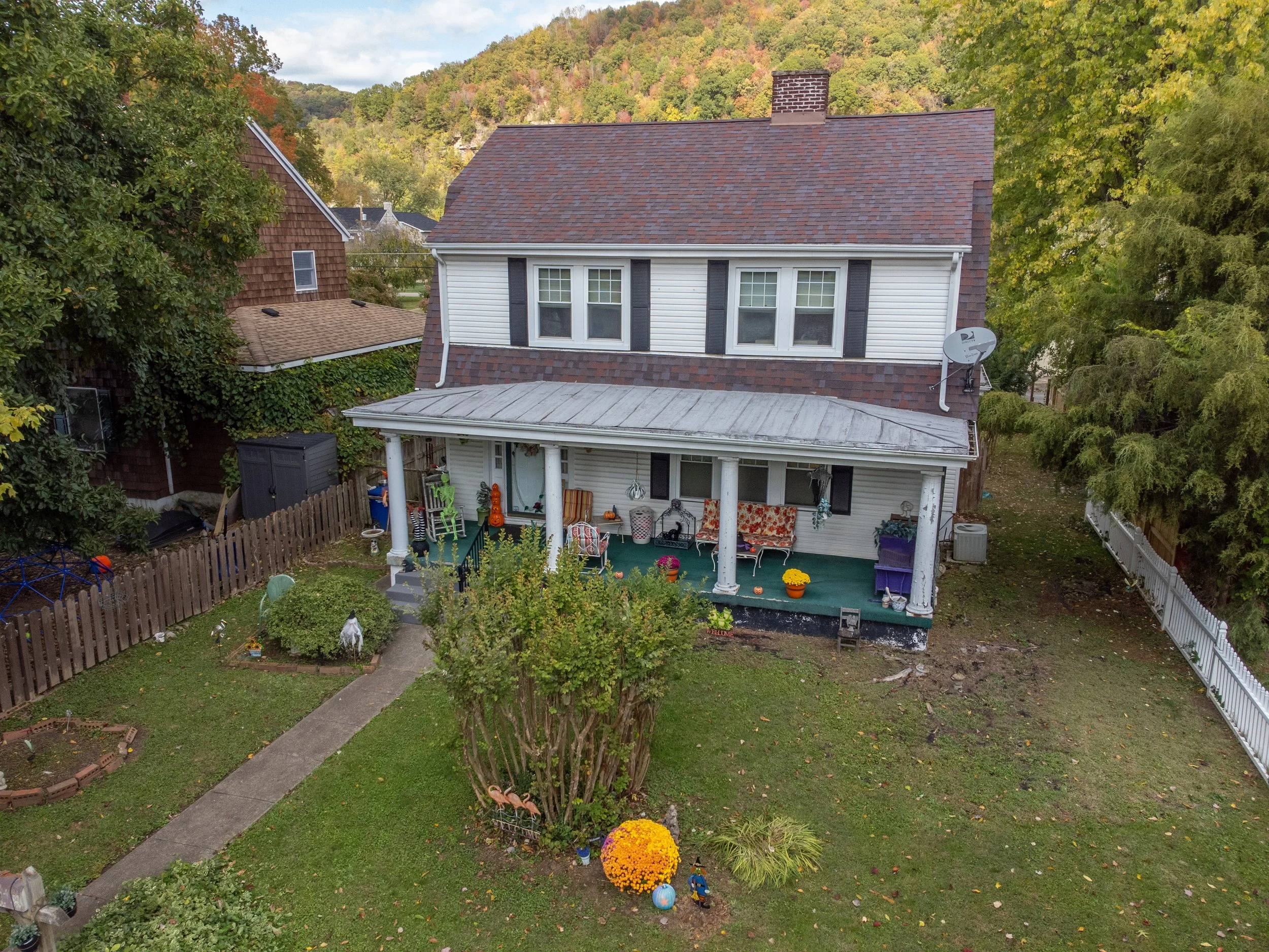 A two-story house with a front porch decorated with various chairs, plants, and Halloween decorations, surrounded by a lawn with a sidewalk, trees, and neighboring houses in the background, during fall.