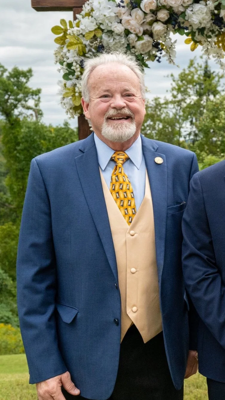 Smiling man in a blue suit and yellow tie, standing outdoors with a background of green foliage and floral arch.