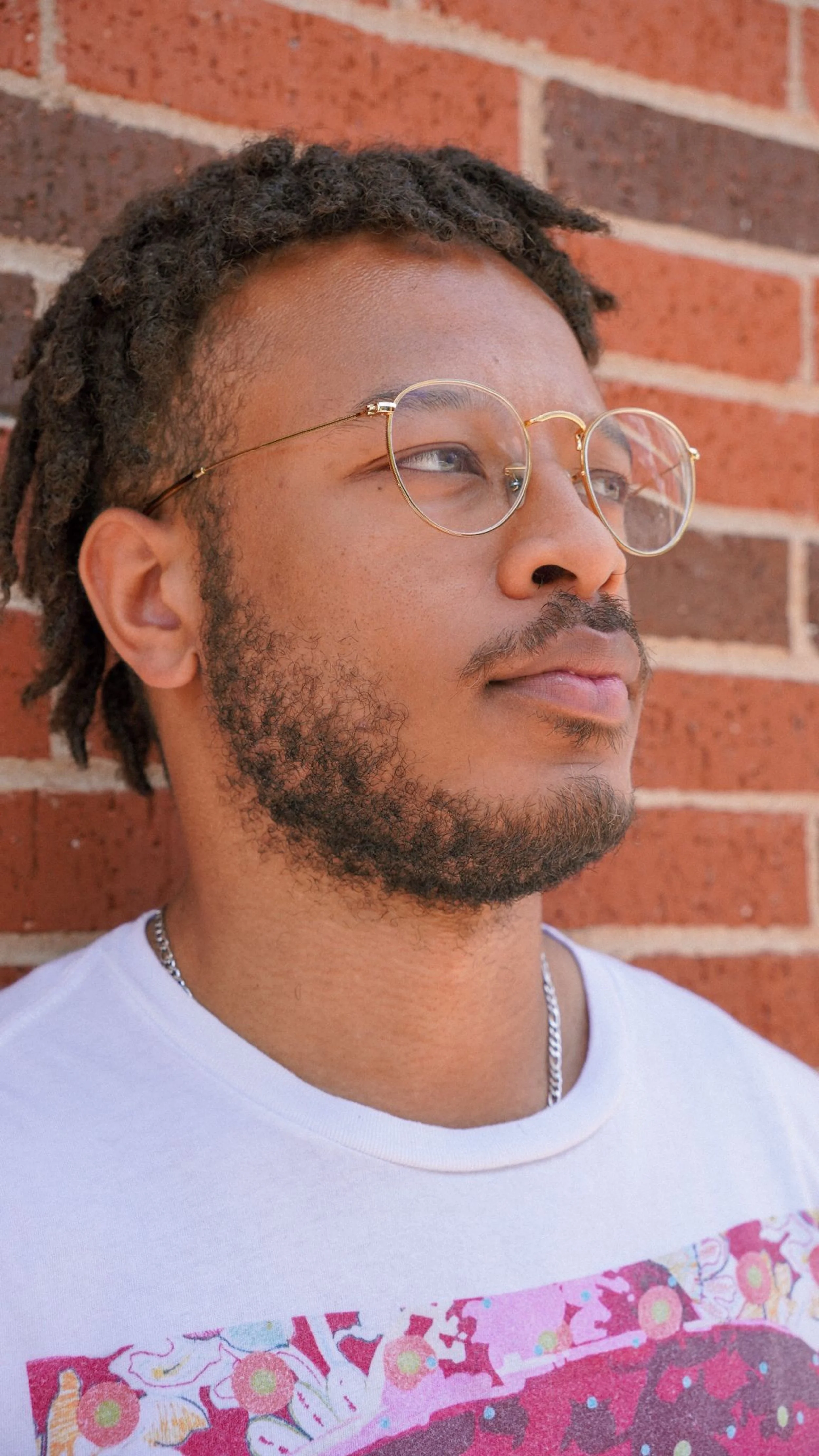 A black male poet in glasses leans against a brick wall outside. He is one of the facilitators for a poetry club.