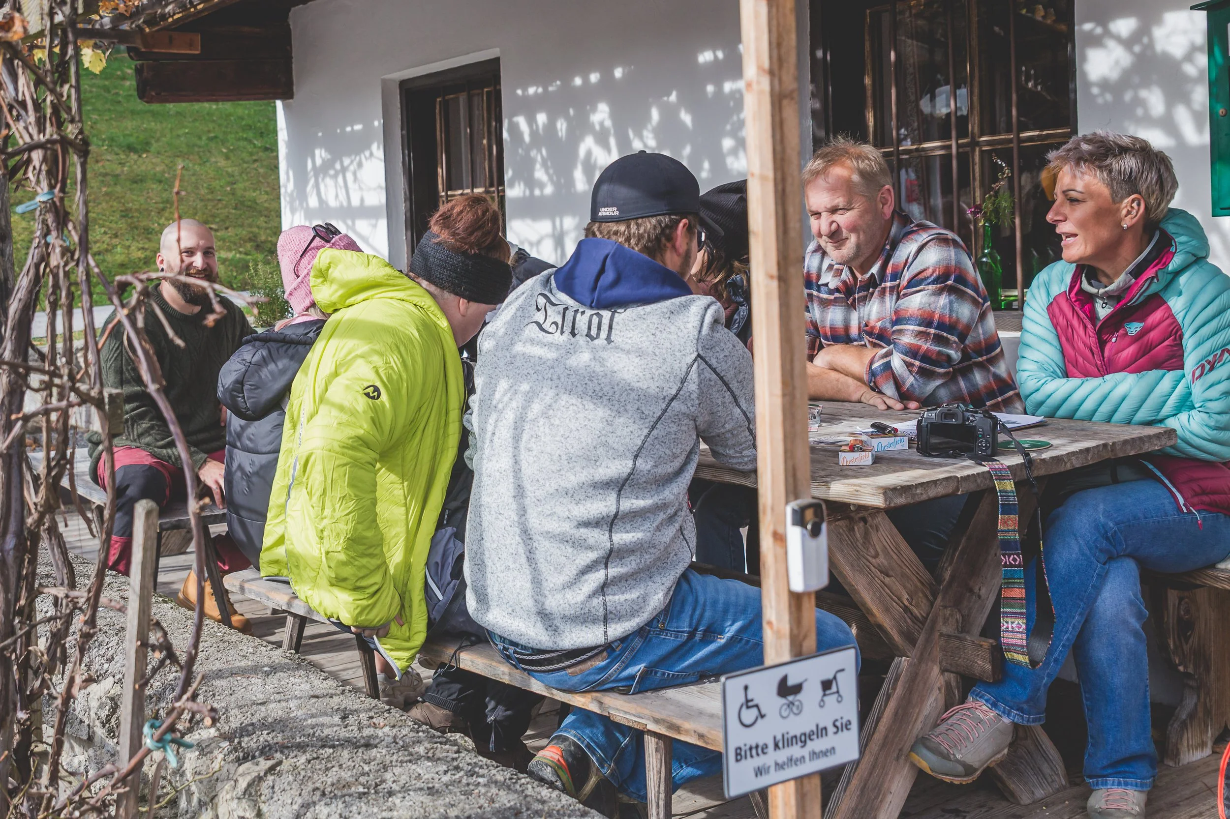 Gruppe von Menschen, die an einem Tisch im Freien sitzen und sich unterhalten, mit einem Schild, das auf Gebärdensprachliche Unterstützung hinweist.
