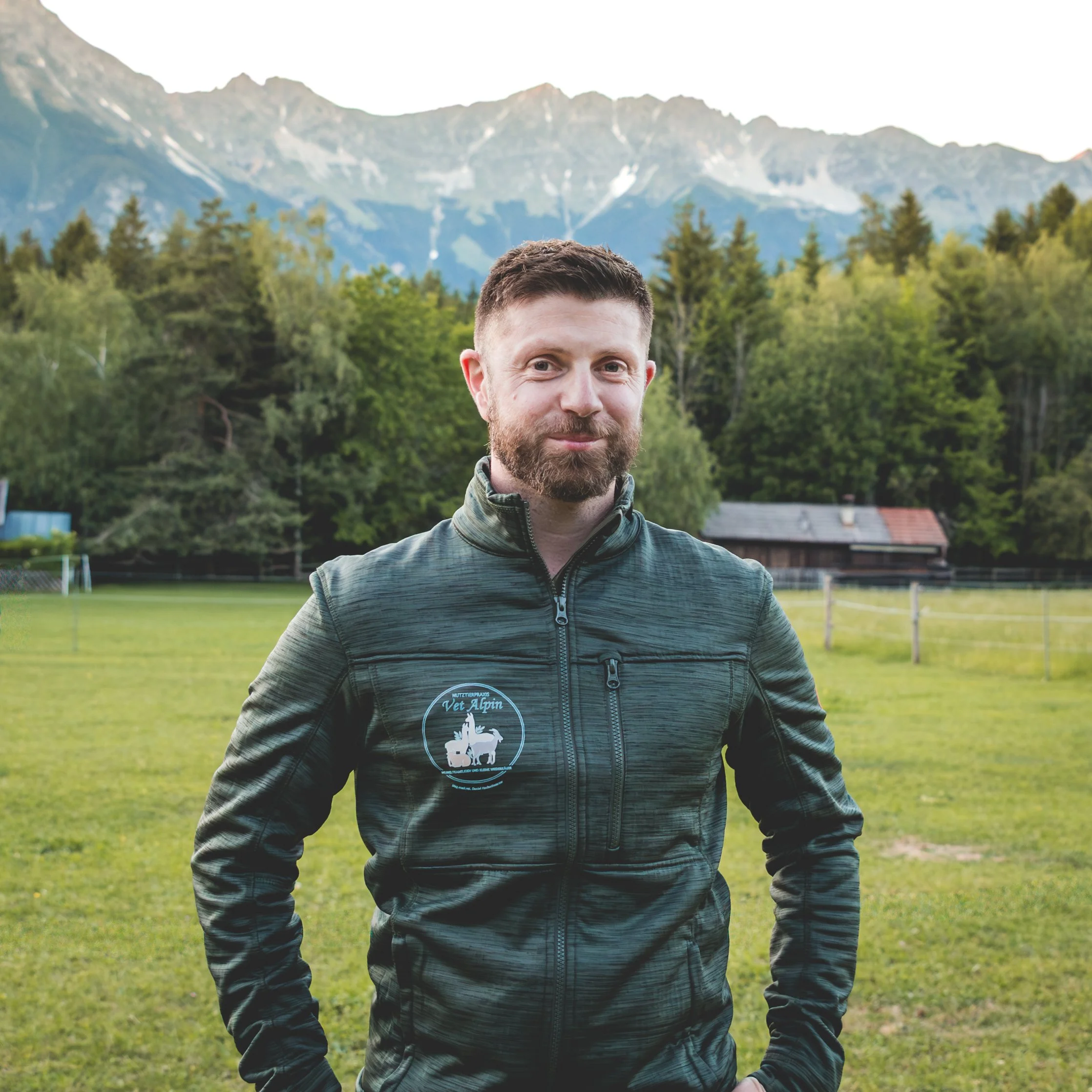 Ein Mann mit Bart und kurzen Haaren steht in einer grünen Wiese mit Bäumen. Im Hintergrund sind Berge und ein kleines Gebäude sichtbar.