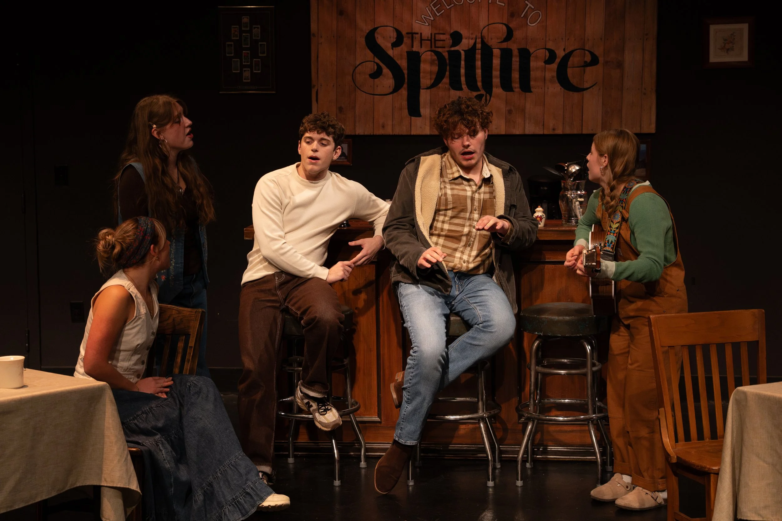 A theatrical performance featuring six young actors on stage at a bar, with three standing and three seated, one of whom is playing a guitar. The background has a wooden sign that reads 'The Spire'.