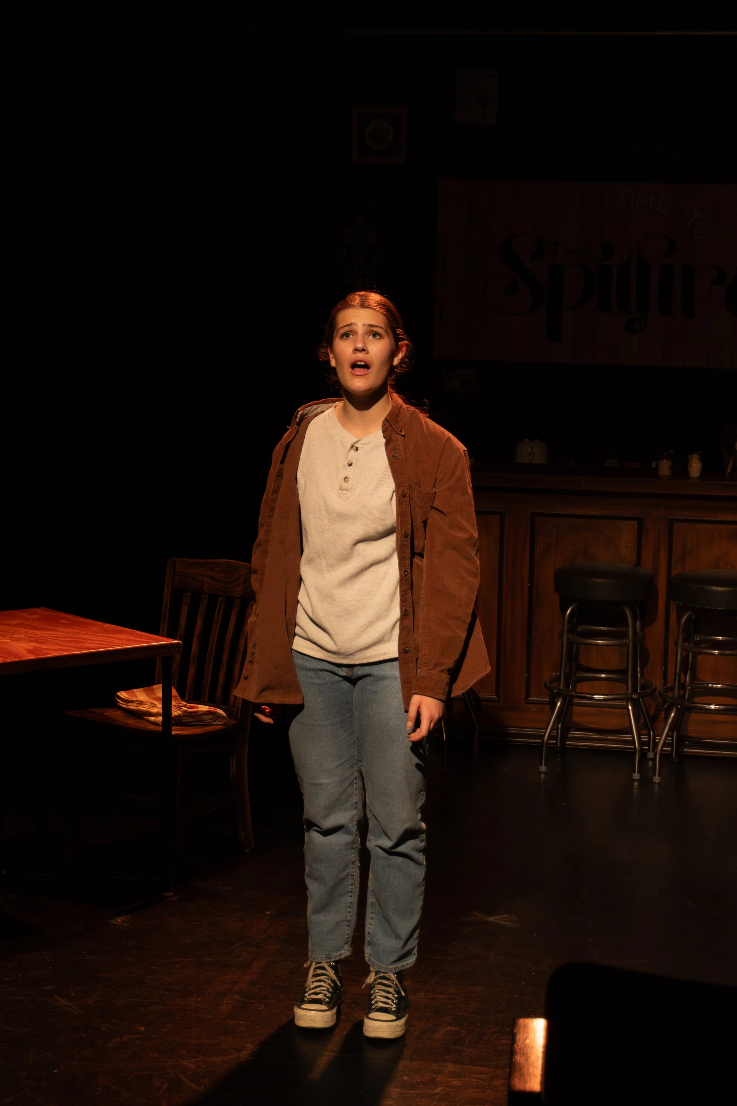 A young woman stands on a dimly lit stage with an expression of surprise or concern, dressed in casual clothing including a beige shirt, brown jacket, jeans, and sneakers. Behind her, there is a wooden bar and stools, and a sign that reads 'Welcome t