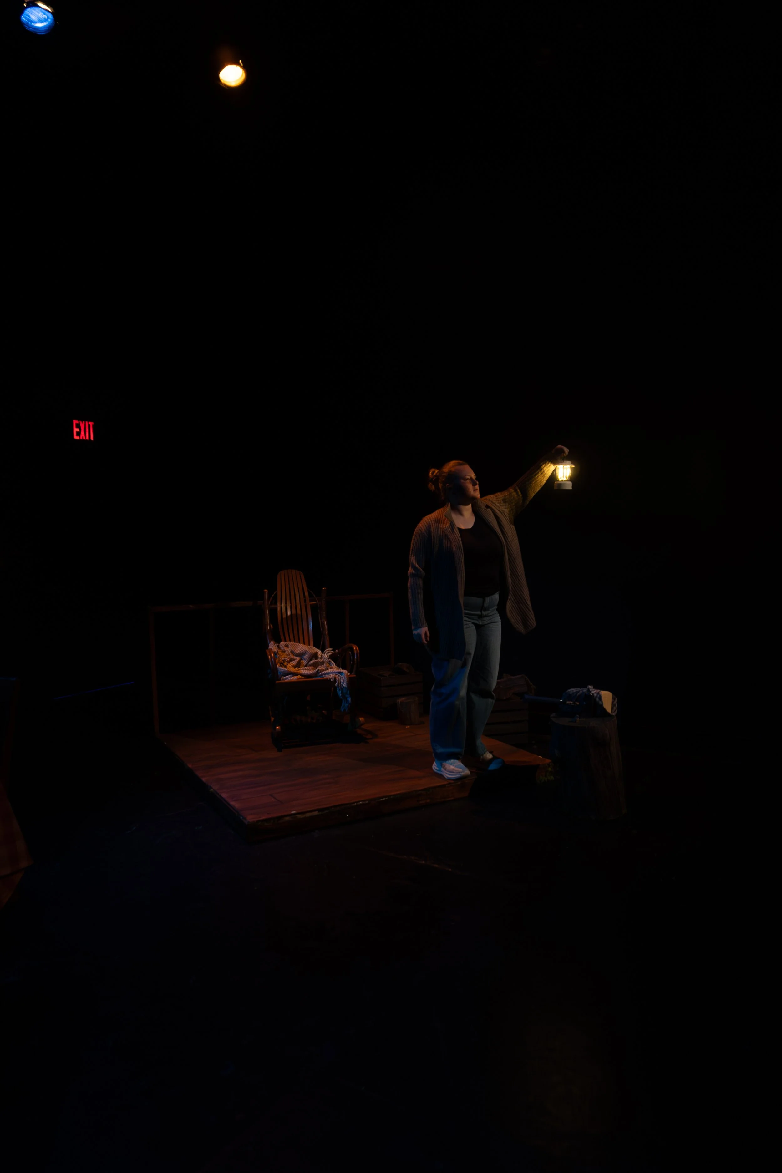 A woman standing on a small stage with a dark background, holding a lantern in her right hand. There is a wooden chair, some boxes, and a small table nearby. An illuminated 'EXIT' sign is visible on the left side.