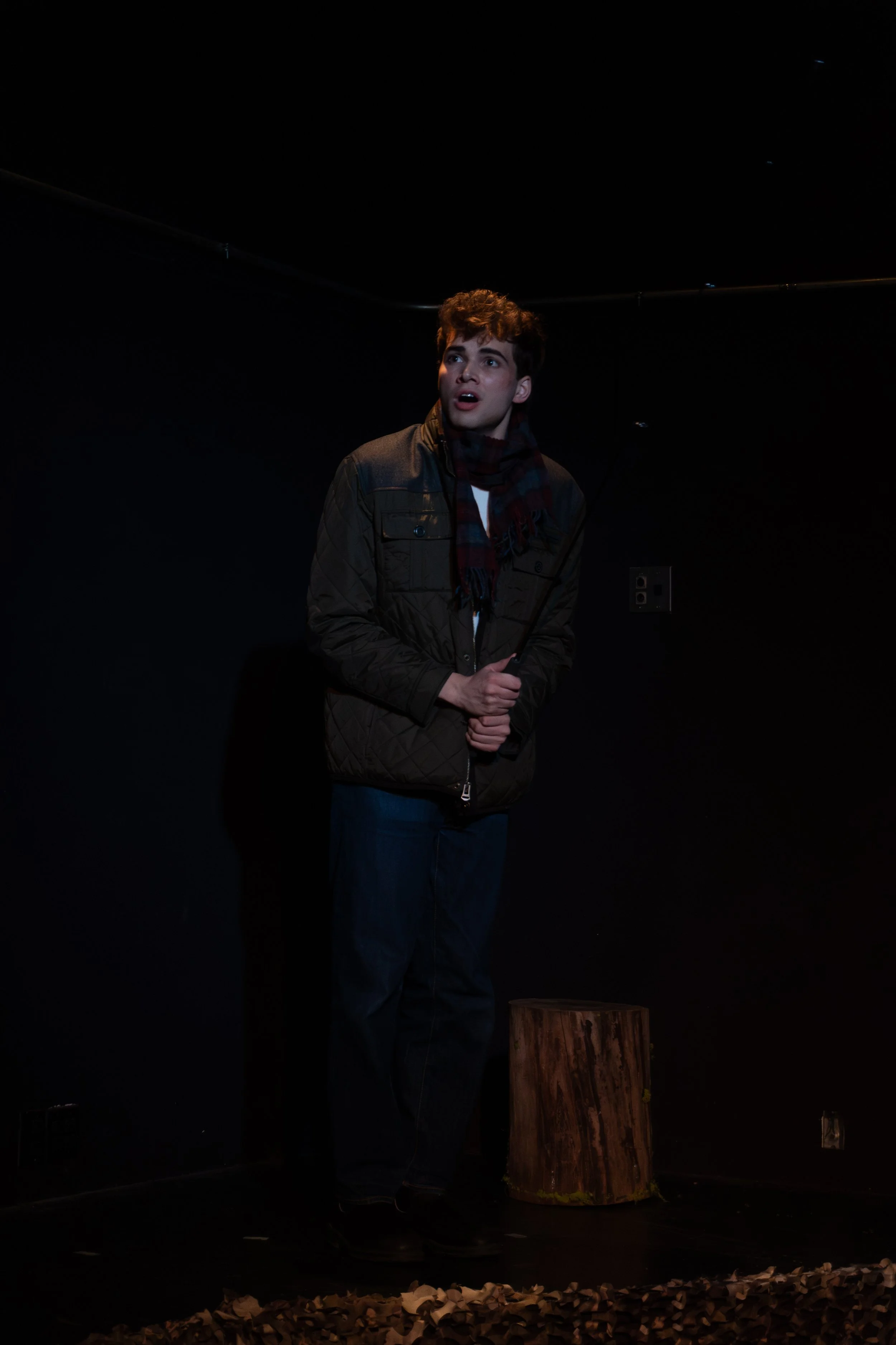 A young man standing in a dark room, looking upward with a surprised or amazed expression, wearing a brown jacket and scarf.
