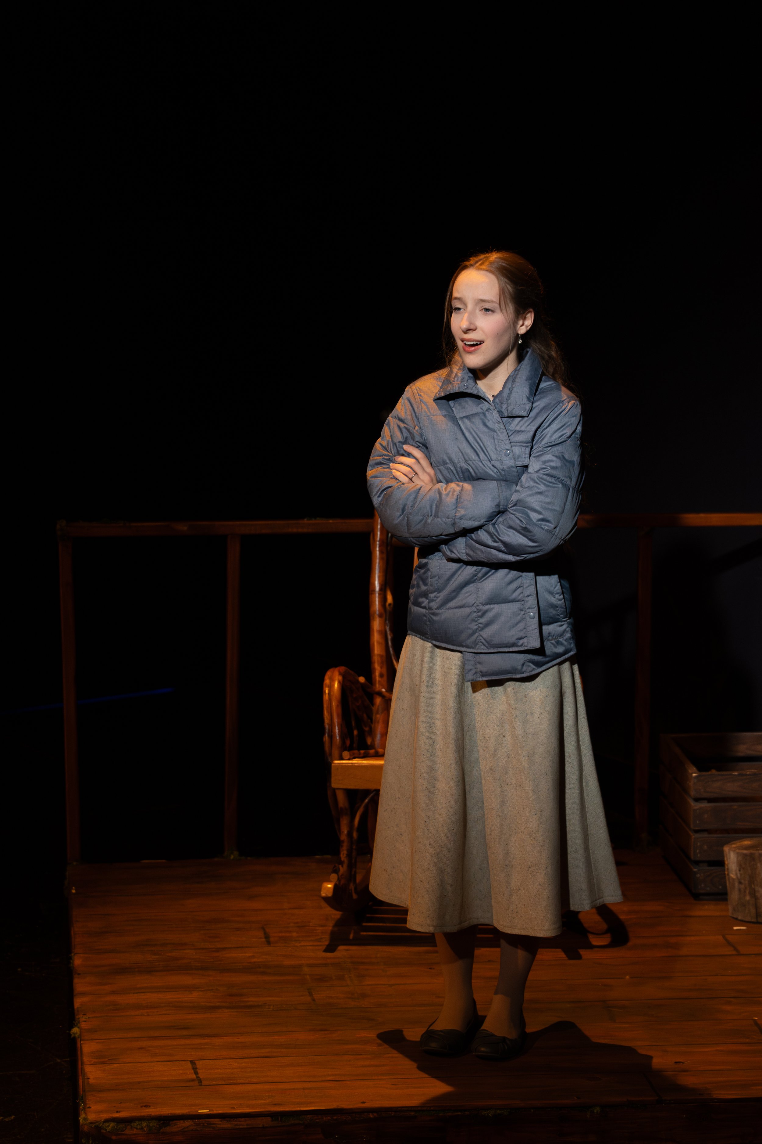 A young woman dressed in period clothing, including a long skirt, a gray jacket, and black shoes, stands on a wooden stage with her arms crossed, appearing to be acting or performing in a theatrical play.