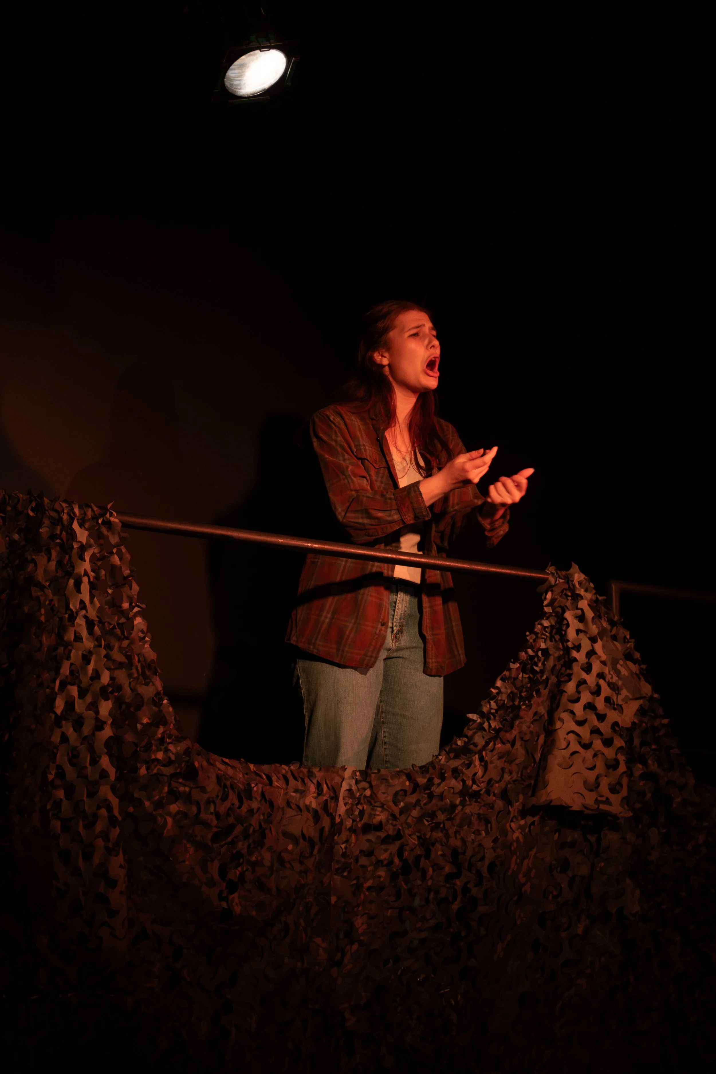 A woman singing or speaking passionately on stage with a microphone in her hand, standing behind a camouflage-patterned barrier or set piece, under a spotlight.