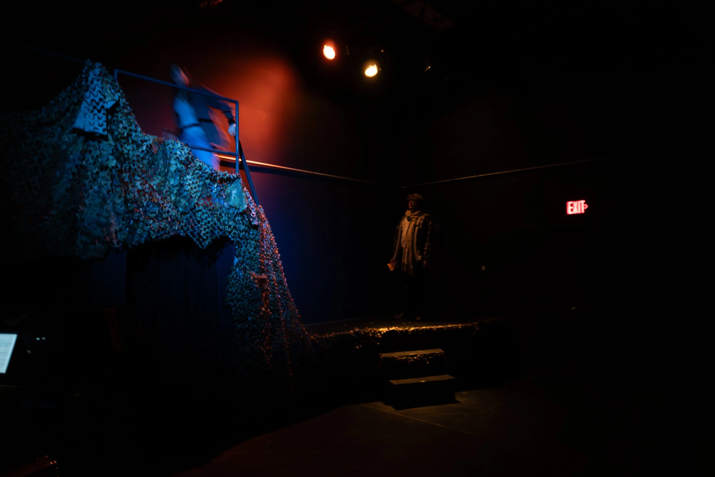 Dimly lit theater stage with a person standing in the background, a person in motion on the stage, blue and red lighting, and a textured island-like structure with multicolored elements in the foreground.