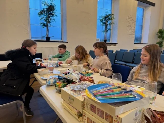 Children sitting at a table with snacks and craft supplies in a bright room with large windows and potted plants.