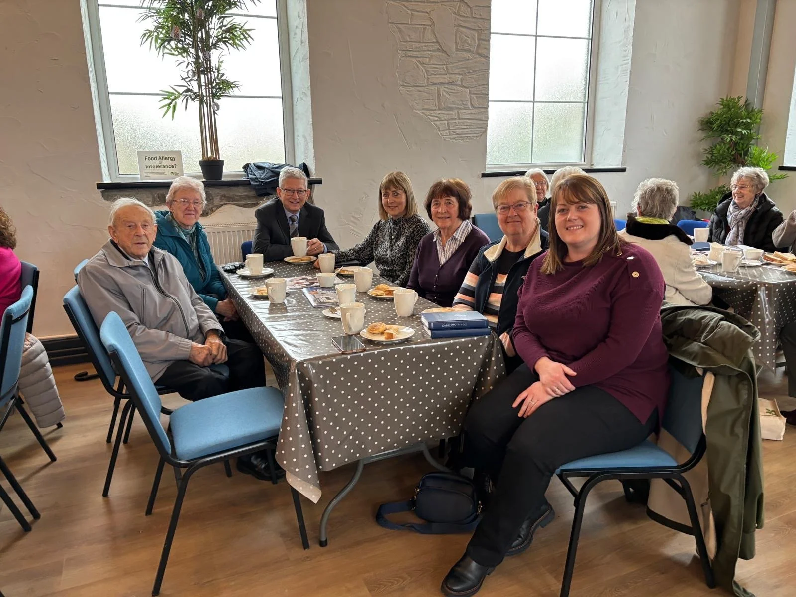 Group of seniors sitting around a table with coffee cups, cookies, and pamphlets, in a well-lit room with large windows and potted plants.