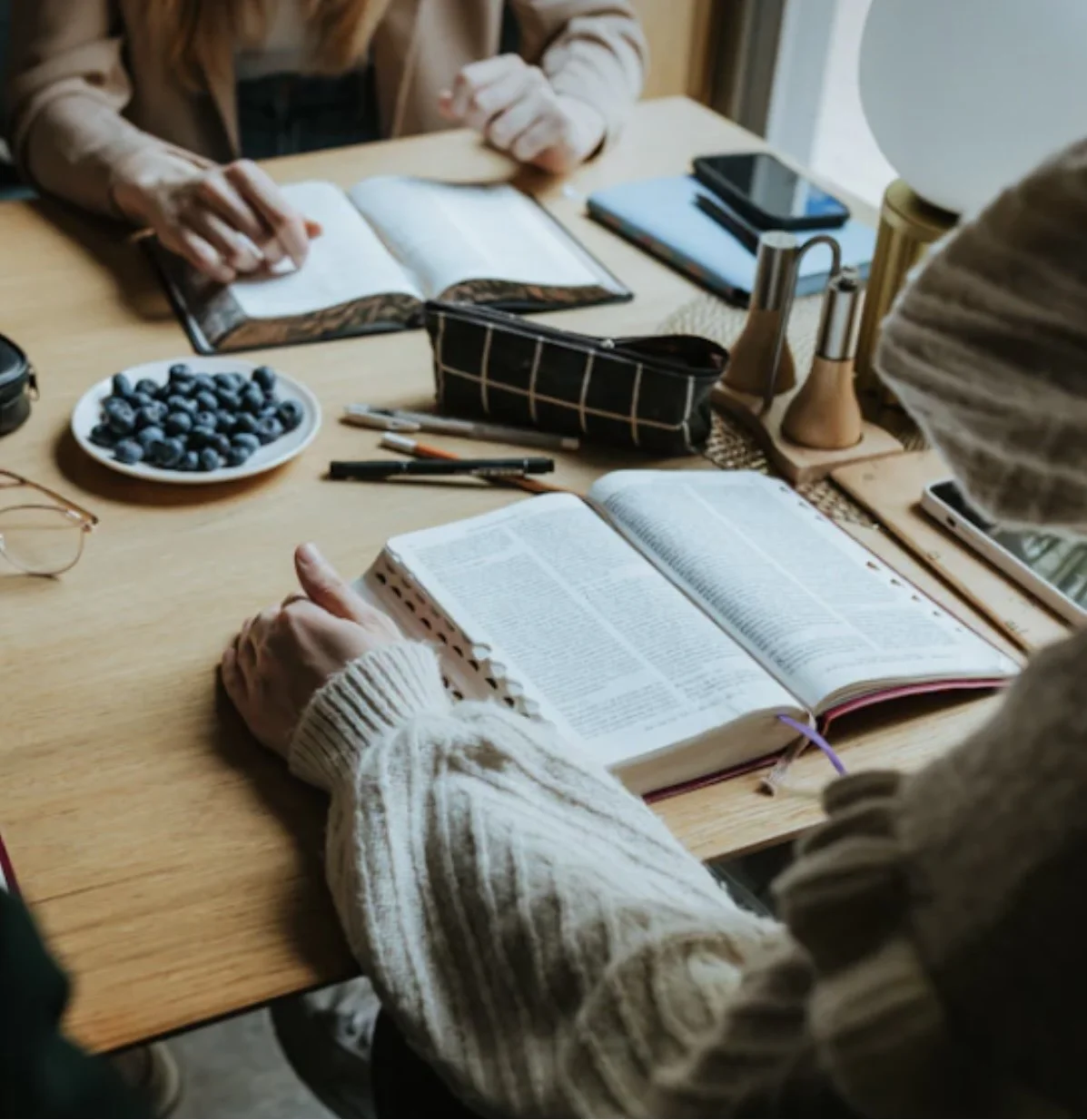 People reading books at a wooden table with notebooks, pens, a plate of blueberries, and electronic devices.