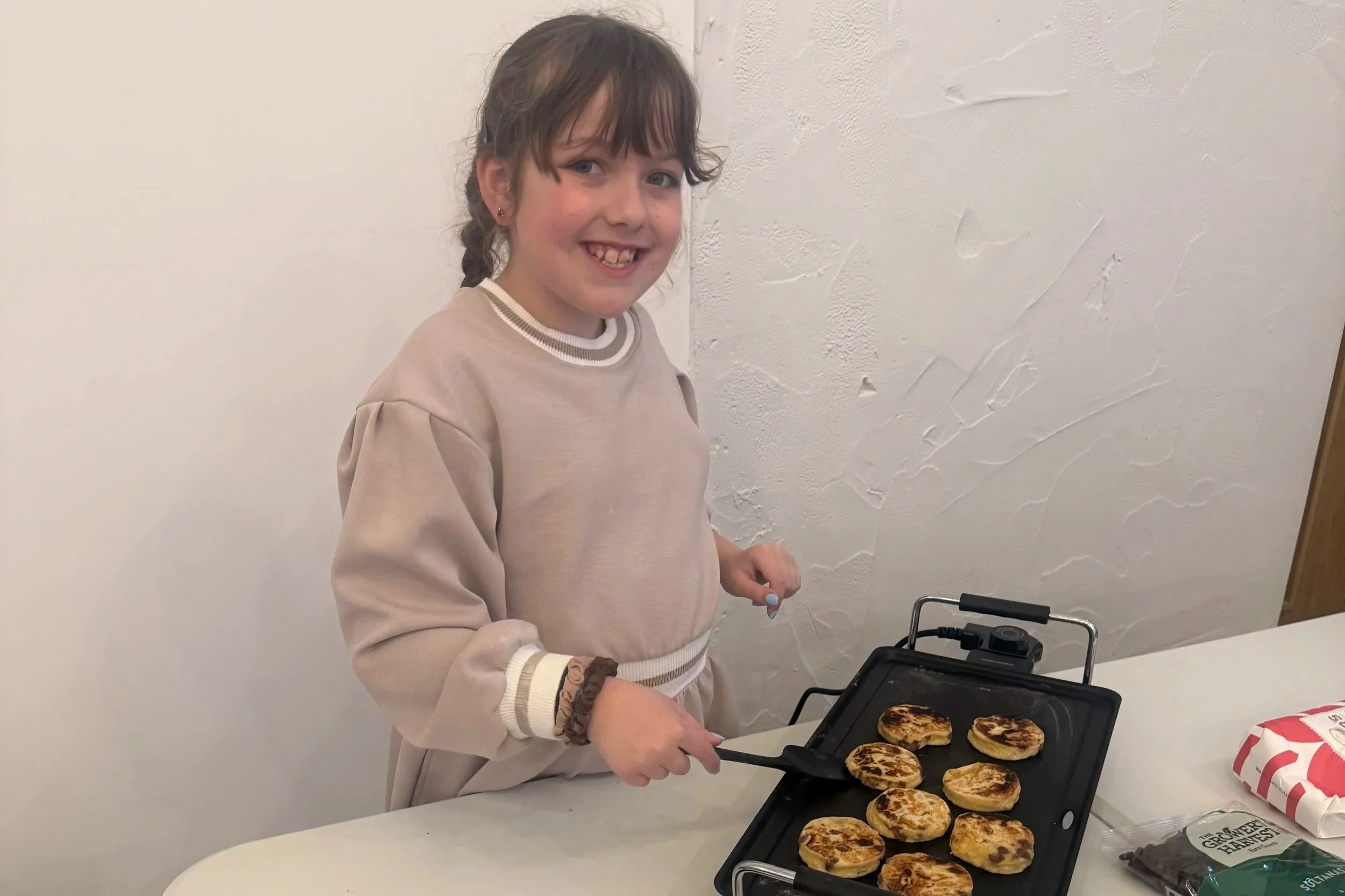 A young girl with brown hair and a beige sweatshirt is smiling while cooking small pancakes on a griddle.