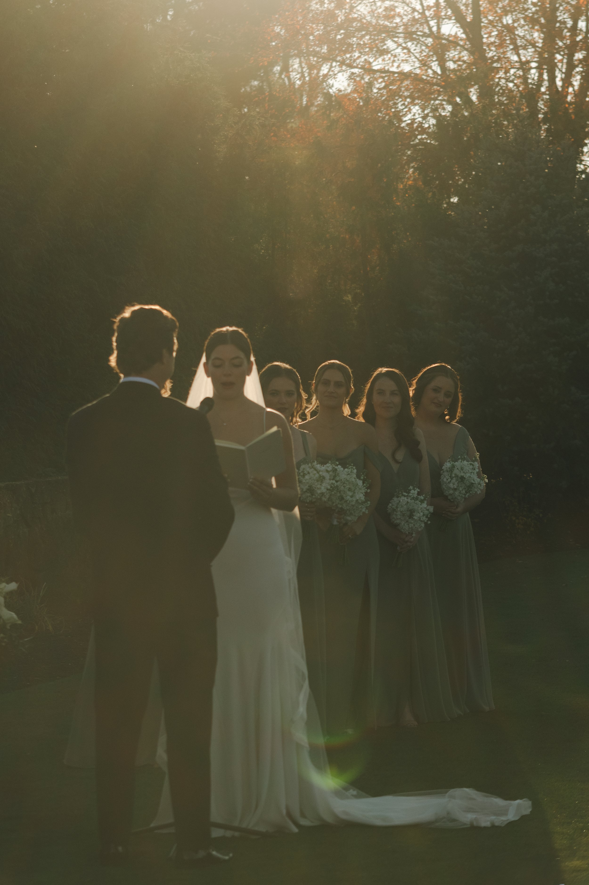 A bride and groom exchange vows outdoors in the late afternoon, with the bride and four bridesmaids holding floral bouquets and standing under the warm sunlight.