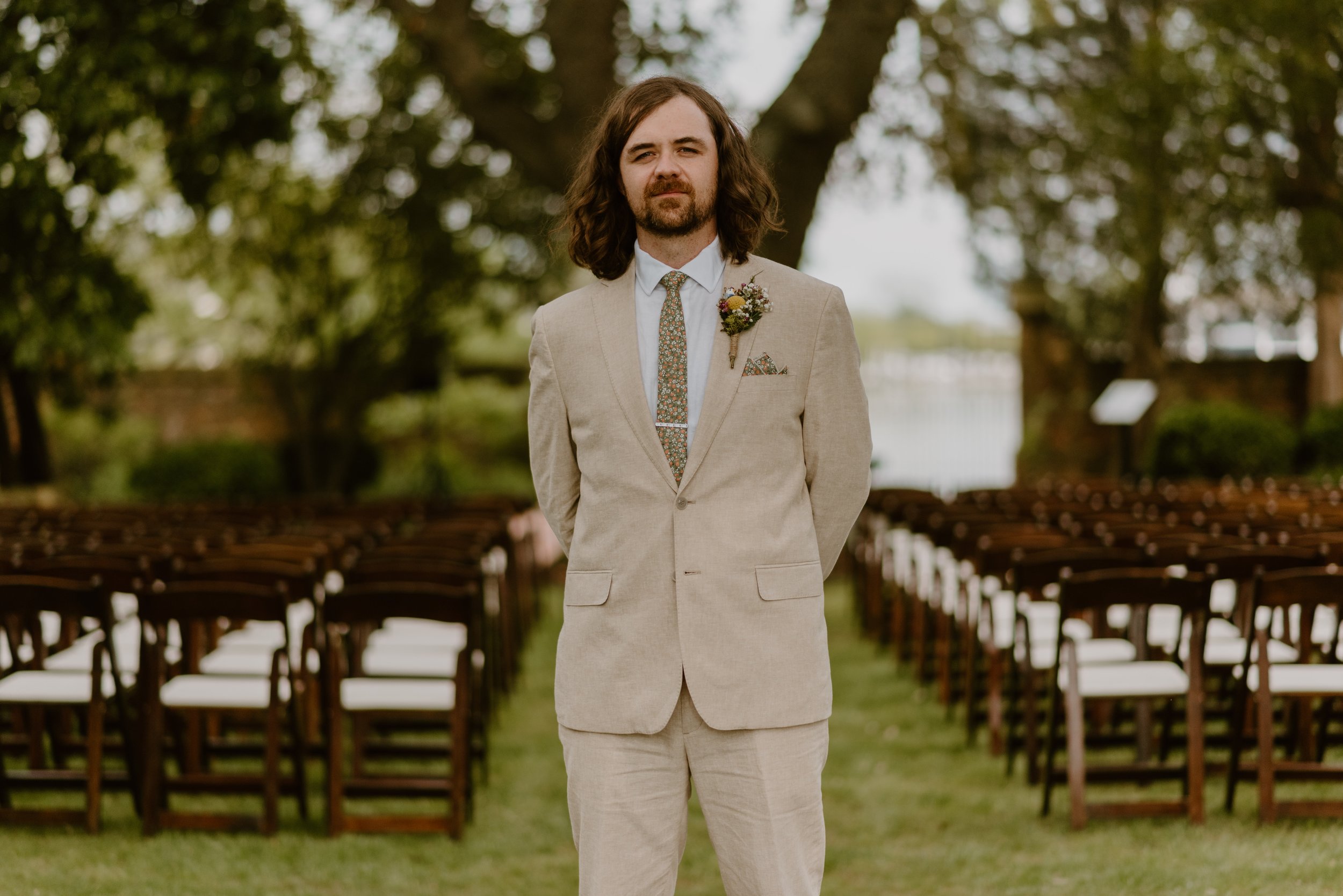 Man in light beige suit with floral tie and flower boutonnière standing outdoors in front of rows of chairs at a wedding venue surrounded by trees.