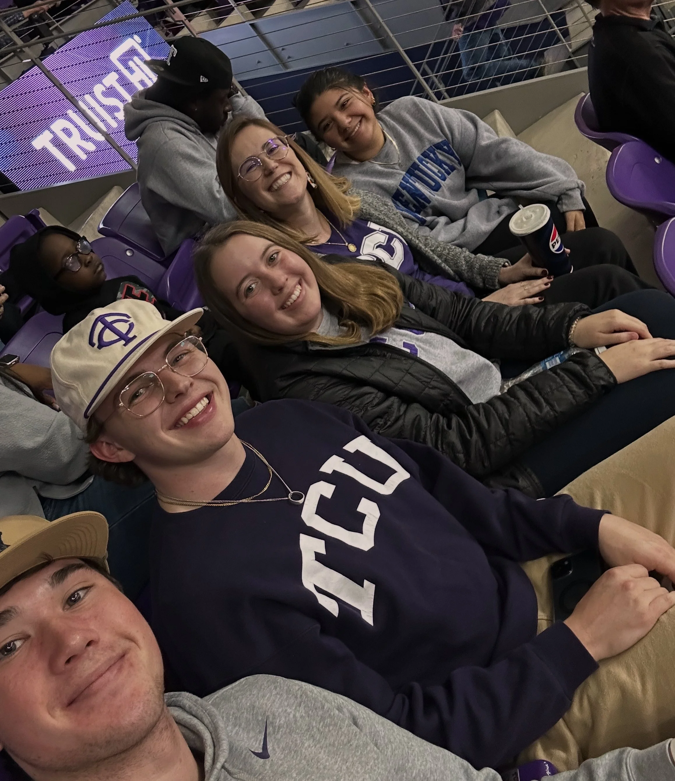 Group of people smiling at a sporting event, wearing TCU and Kentucky apparel, seated in a stadium with purple seats and a "TRUST" sign in the background.