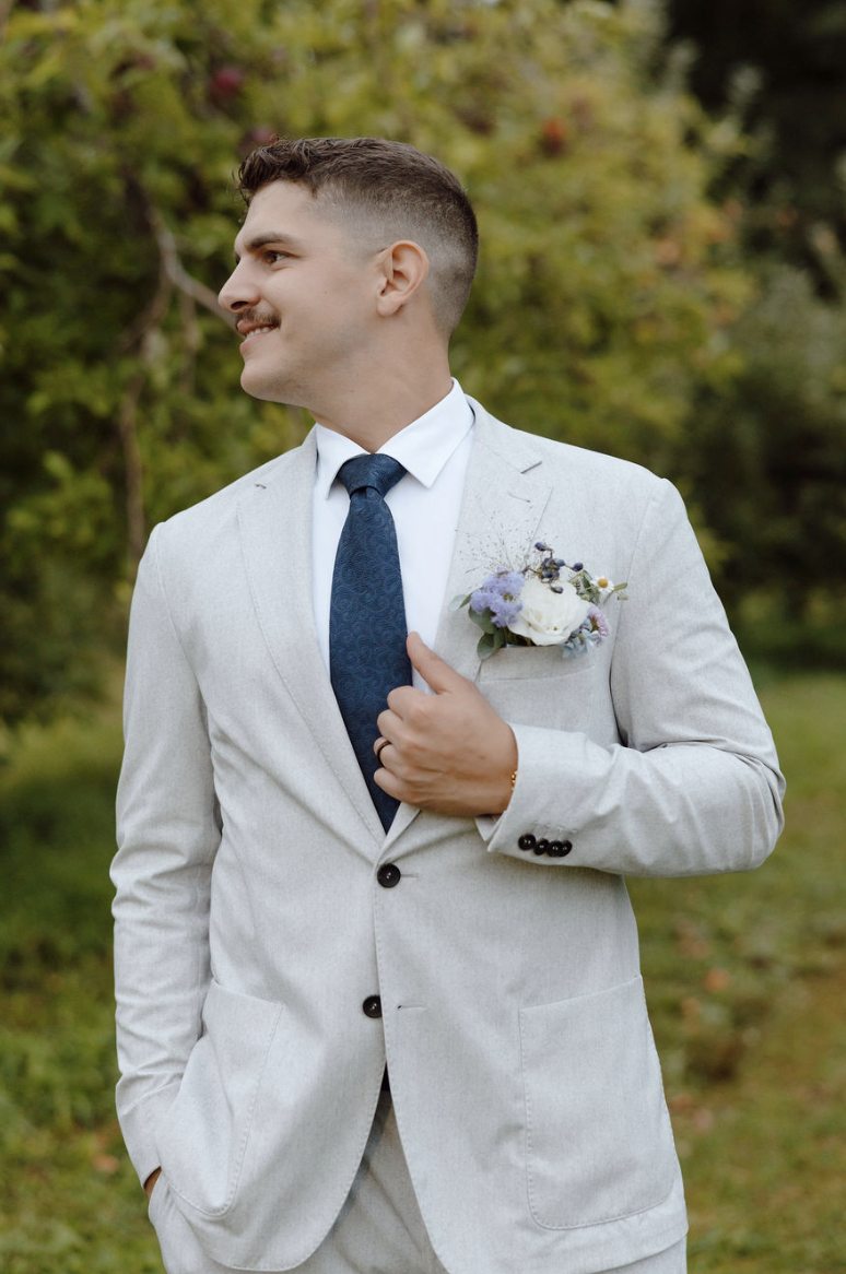 A man in a light grey suit with a white shirt and dark blue tie, standing outdoors near greenery, holding his chest with one hand that has a boutonnière with white and purple flowers.