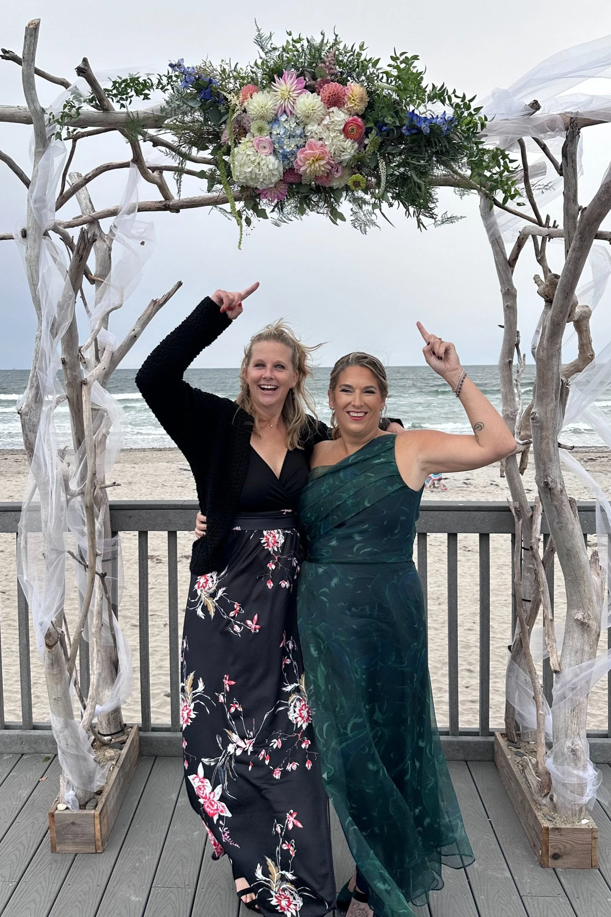 Two women standing under a beachside wedding arch decorated with flowers and driftwood, smiling and raising their arms during a celebration.