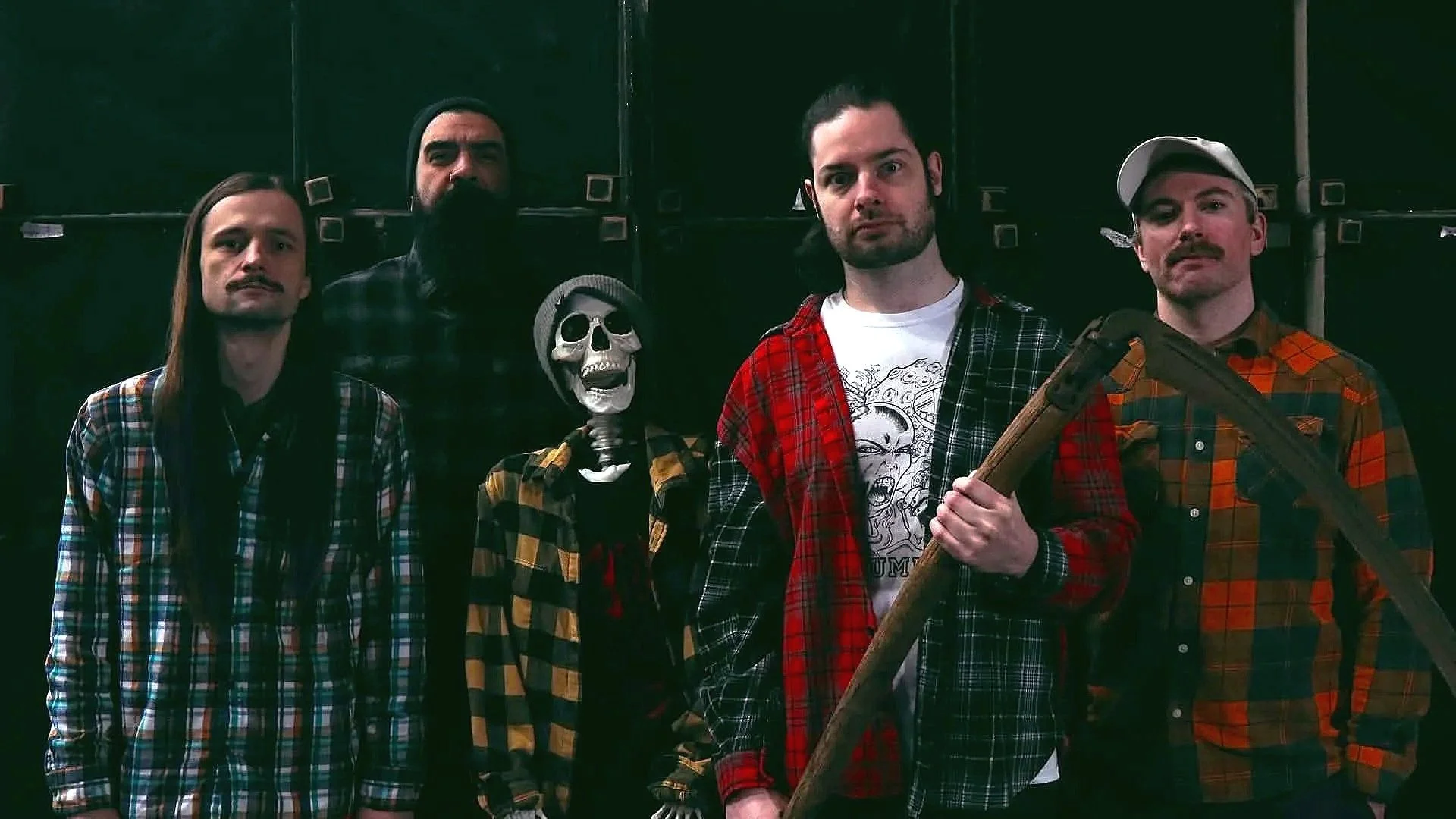 Five men with long hair, beards or mustaches wearing plaid shirts, standing against a dark background, one holding a wooden mannequin with a skull face.