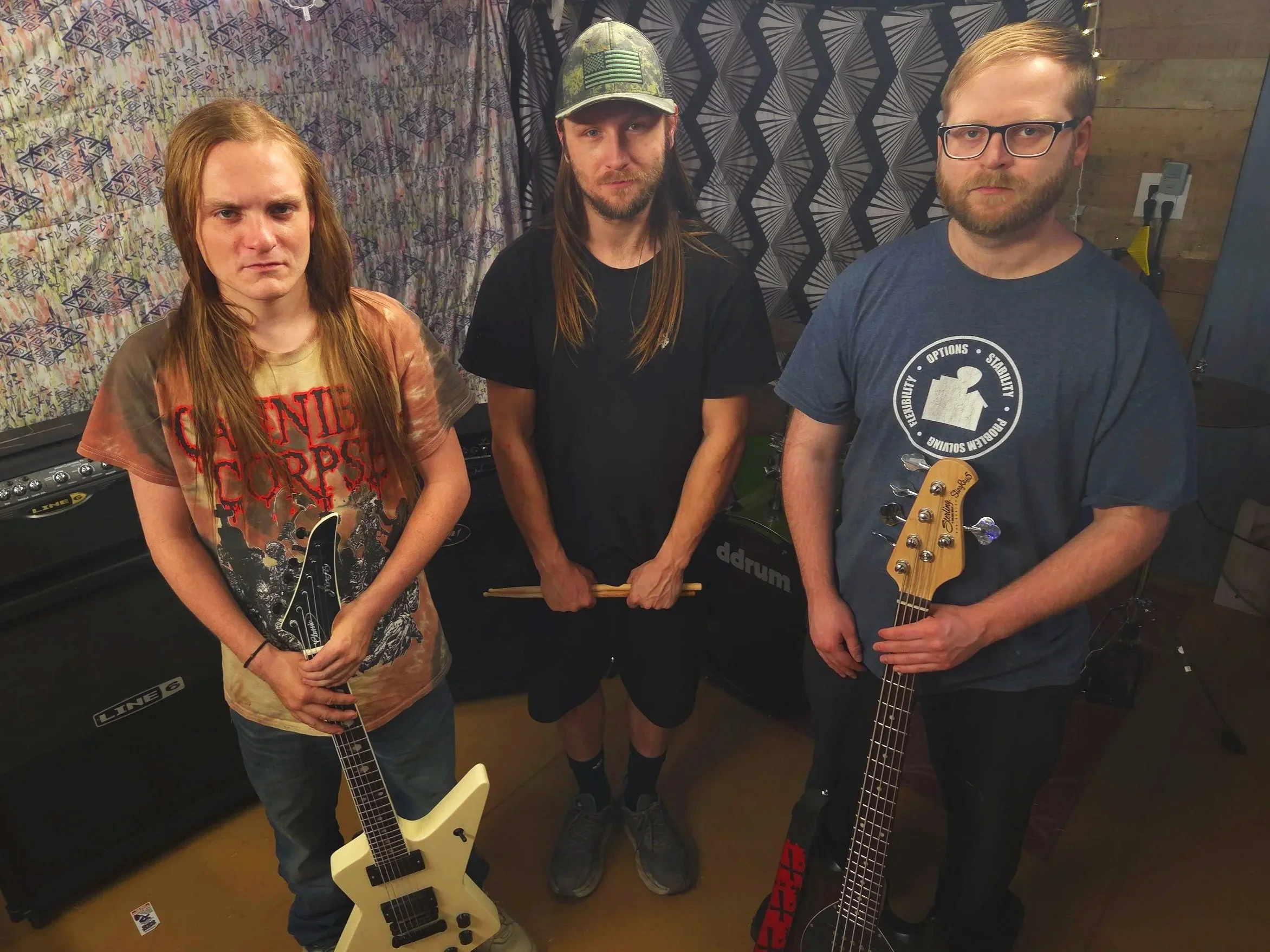 Three men standing indoors, two holding guitars, one holding drumsticks, with musical equipment in the background.
