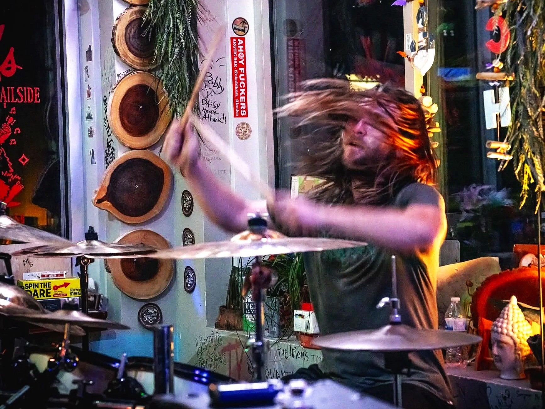 A drummer with long hair playing drums in a colorful room with various decorations and artwork.