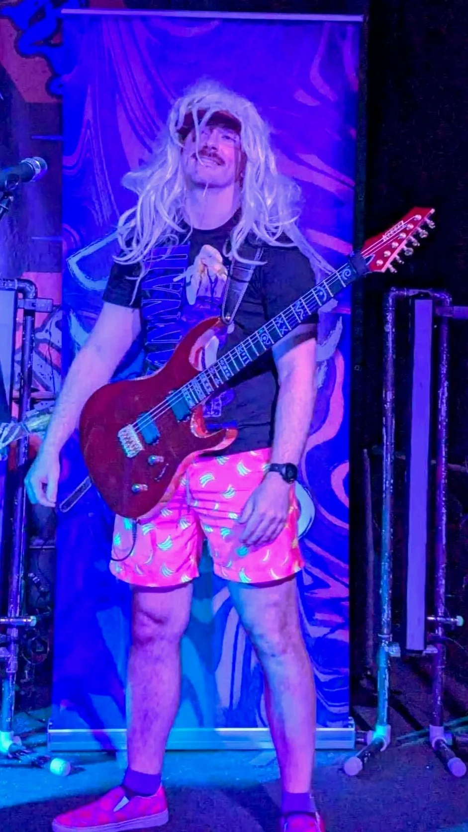 A man with long blonde hair and a mustache holding a red electric guitar on stage, wearing a black graphic T-shirt, pink shorts with a banana pattern, pink shoes, and a black watch, with artsy purple and blue stage lighting.