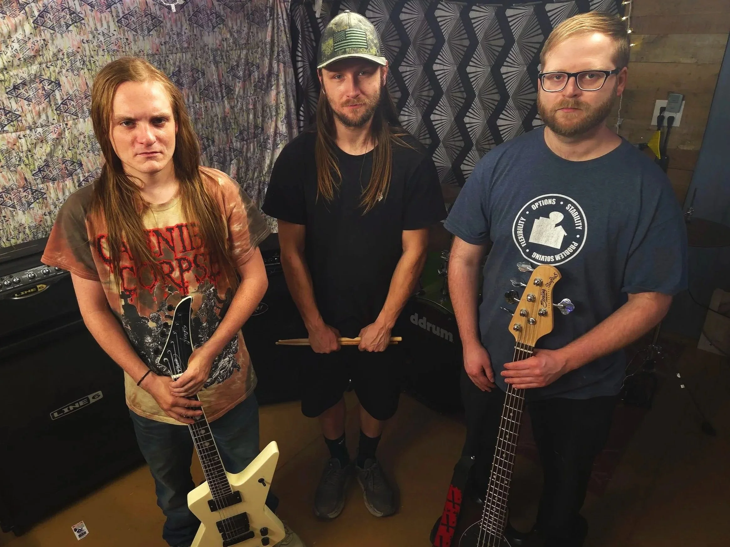 Three musicians holding guitars and drumsticks in a music studio with patterned wallpaper and equipment in the background.