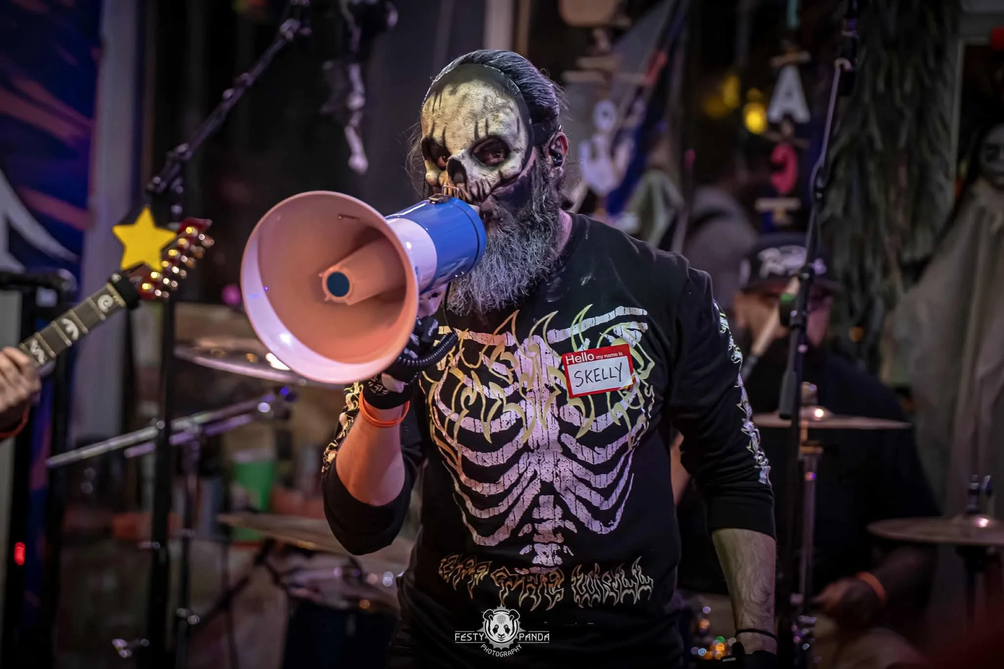 A man with skeleton makeup on his face, beard, and hair holding a loudspeaker to his mouth wearing a black shirt with skeleton bones and a name tag that says 'Skelly,' at a Halloween or costume party event.