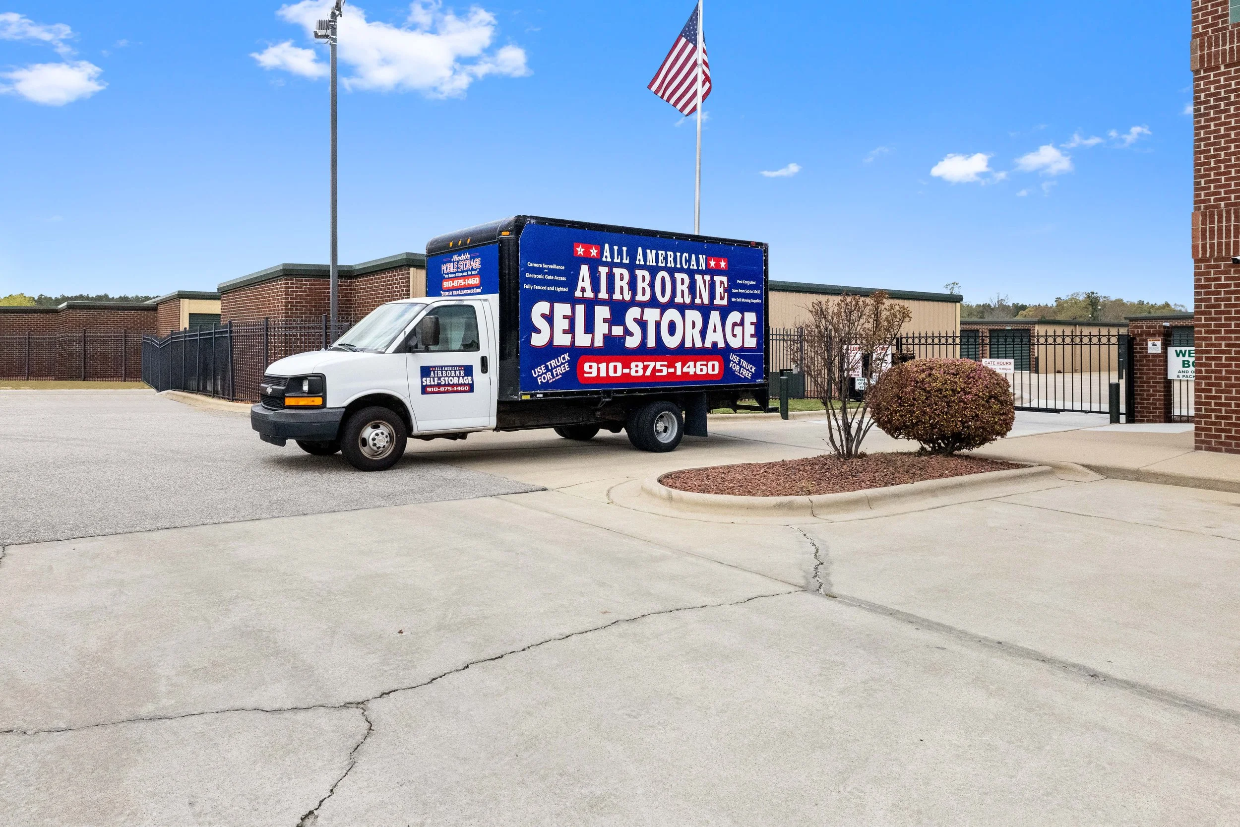A white truck with a large blue advertisement for 'All American Airborne Self-Storage' parked in front of a gated storage facility. The advertisement includes a phone number and mentions free use of the truck. An American flag is flying on a pole beh