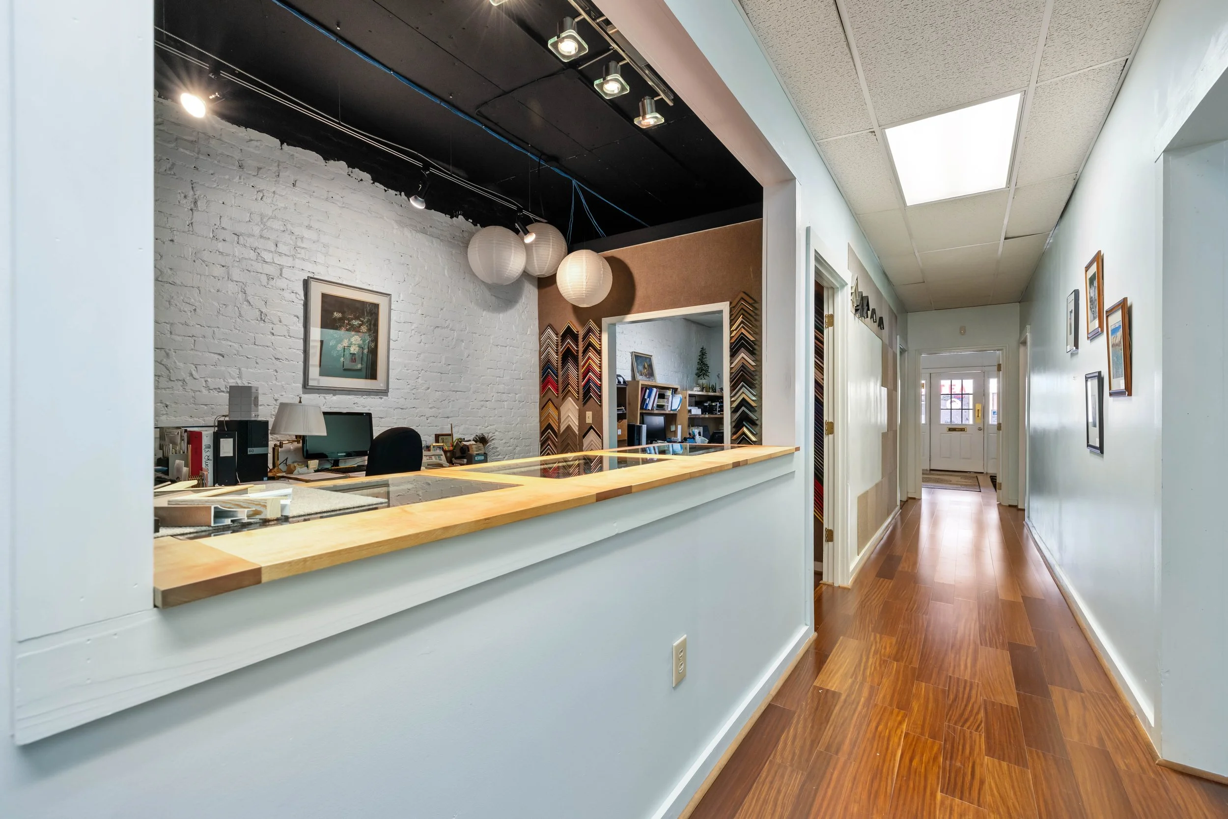 Office reception area with a wooden counter, white brick wall, paintings on the wall, and items on the desk, with a hallway extending into the building.