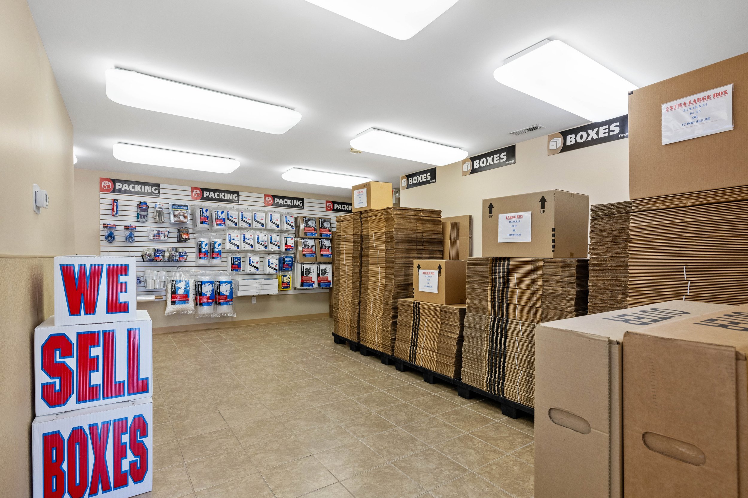 Interior of a packing supplies store with stacks of cardboard boxes on pallets and a wall display of packing accessories. A sign in the foreground reads 'WE SELL BOXES' in large red and blue letters.