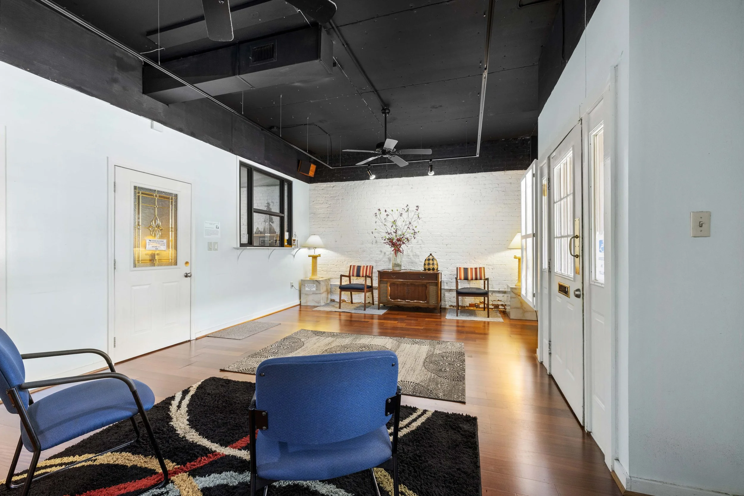 Interior of a waiting room with blue chairs, a black rug, and a white brick wall with a table and chairs in front of it, decorated with a vase of flowers.
