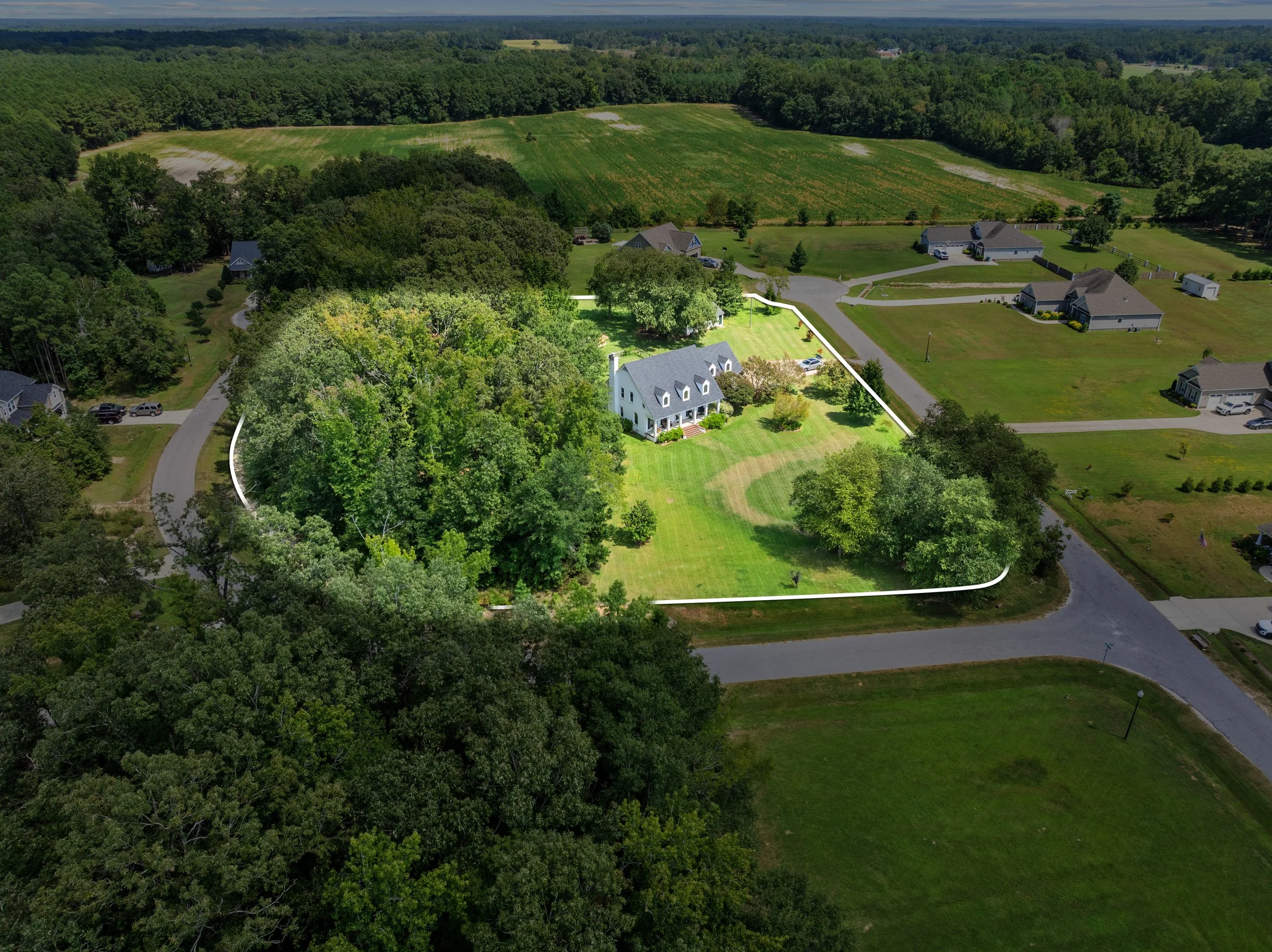Aerial view of a white house surrounded by a large backyard, trees, and nearby houses in a suburban neighborhood.