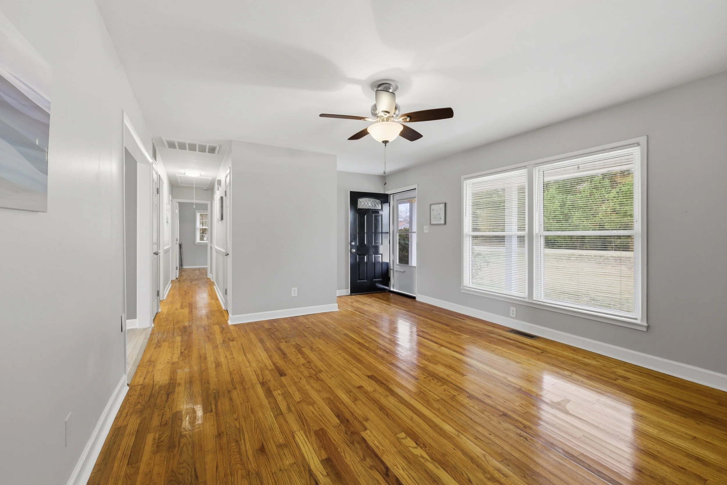 Empty living room with hardwood floors, large windows, a ceiling fan, and a black front door.
