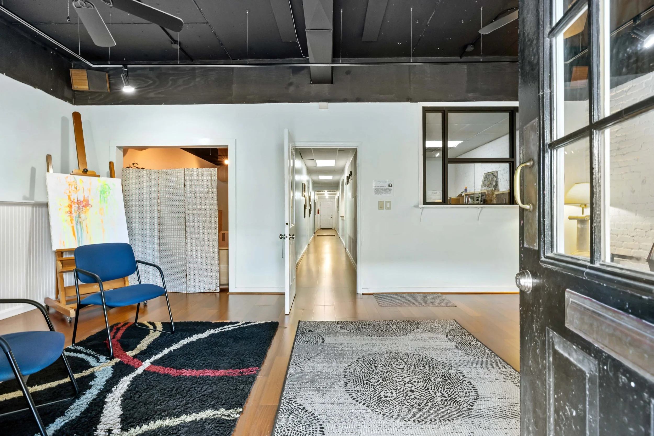 Indoor view of an art gallery with chairs, paintings, and rugs, seen through an open black door with a brass handle.