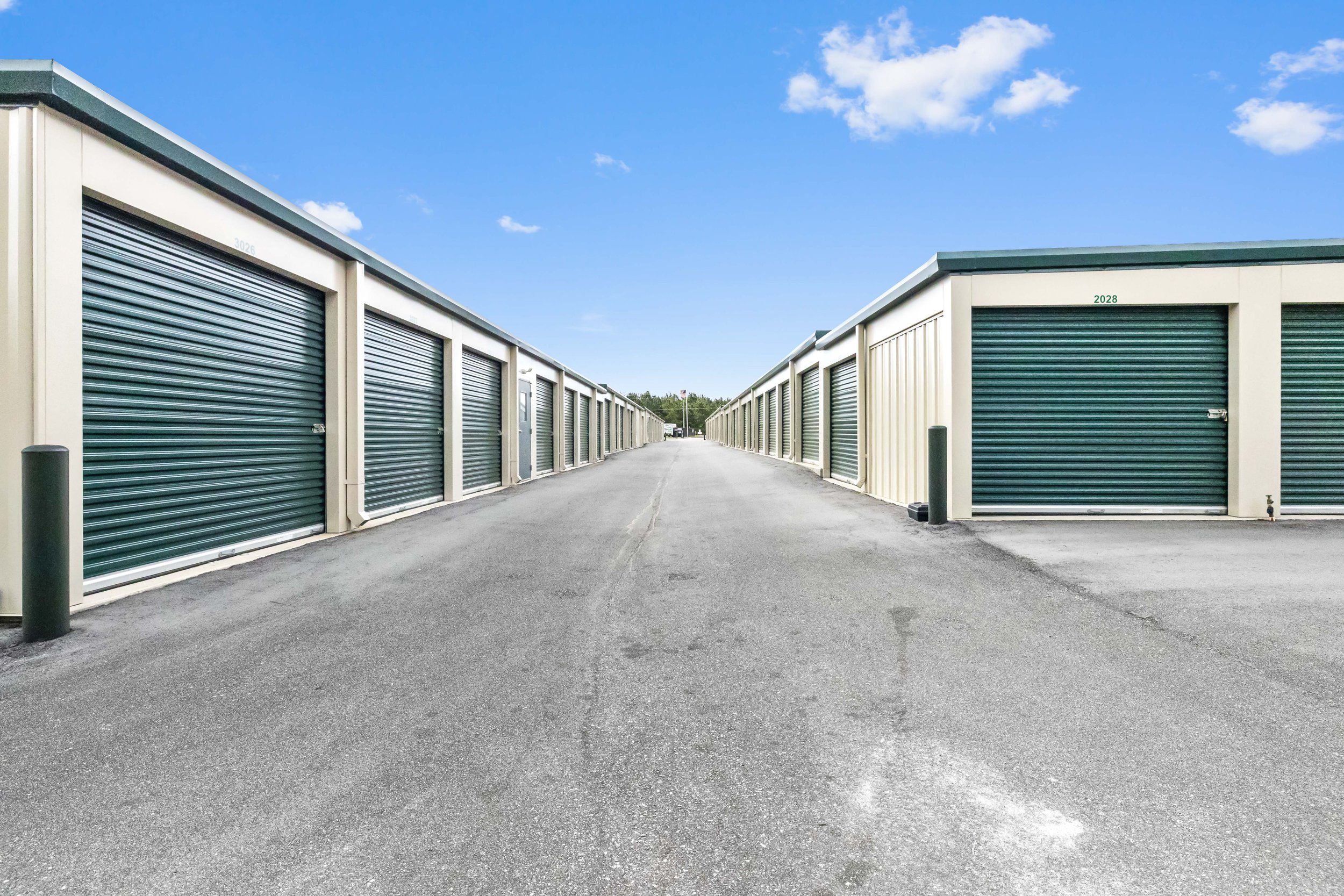 A row of storage units with roll-up doors on either side of a paved driveway, under a blue sky with a few clouds.