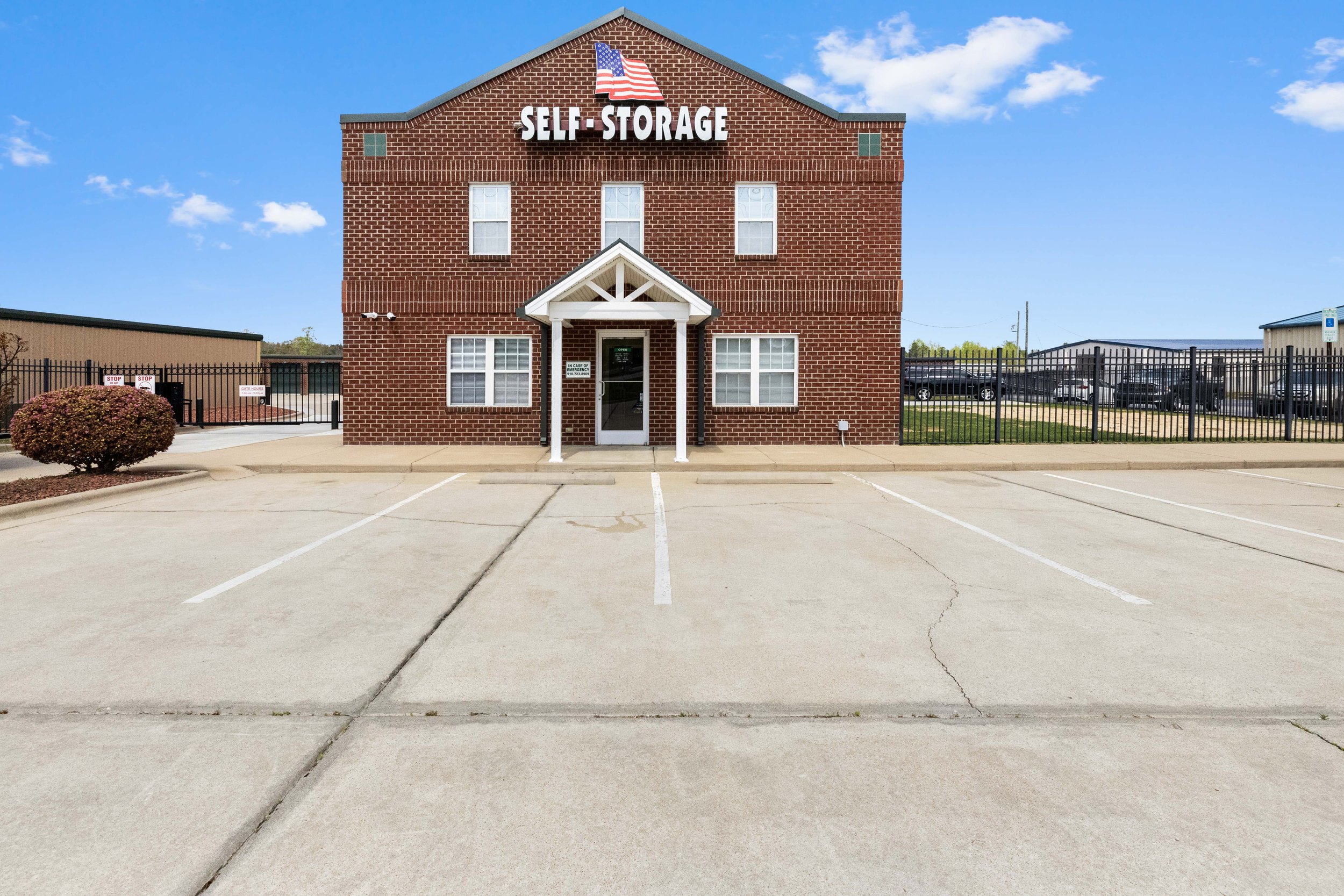 A brick building with a sign that reads 'Self-Storage' and an American flag on top. There is a small entrance with a porch, and parking spaces in front. The sky is blue with some white clouds.