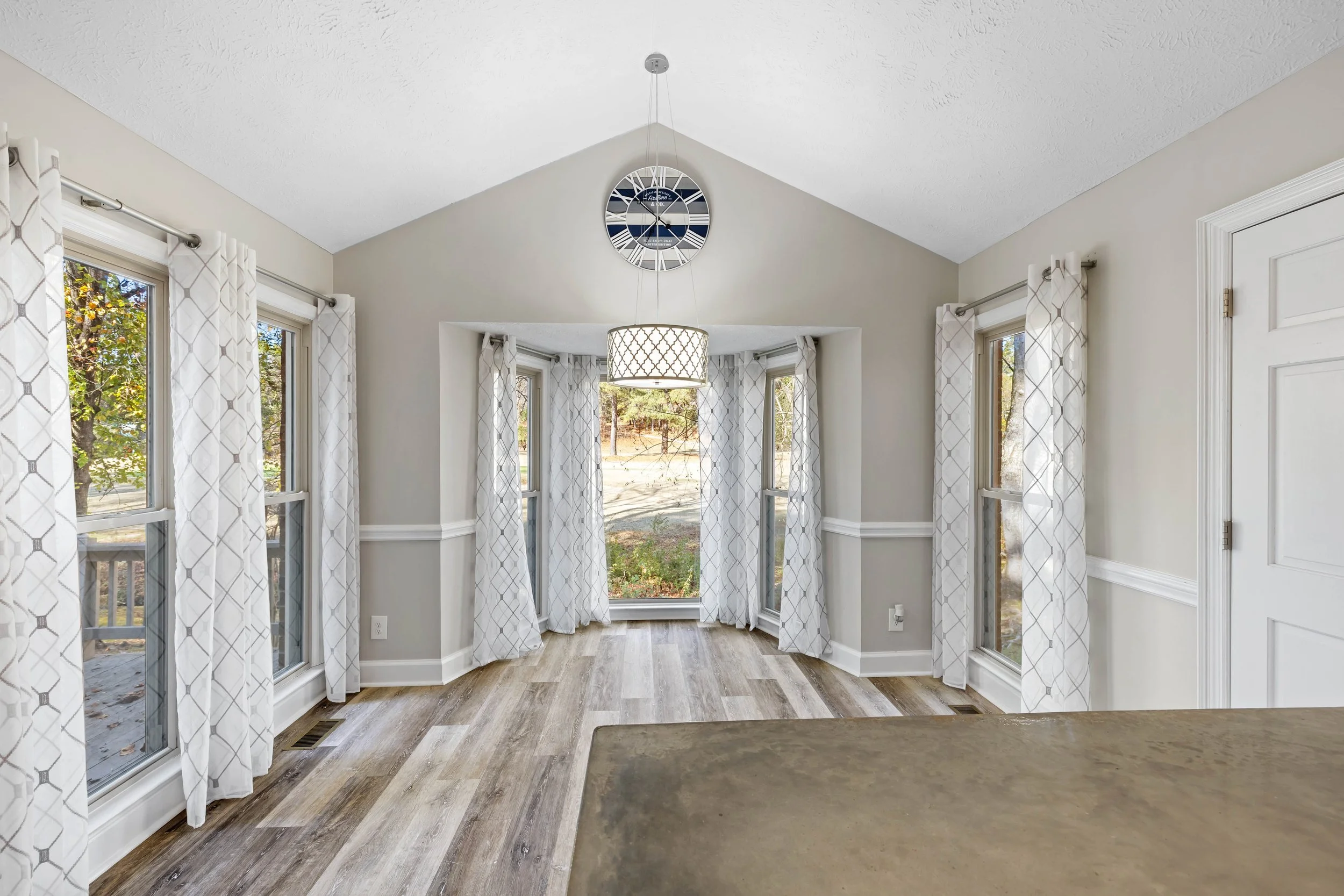 Bright dining area with multiple large windows, white patterned curtains, a modern hanging light fixture, and a wall clock