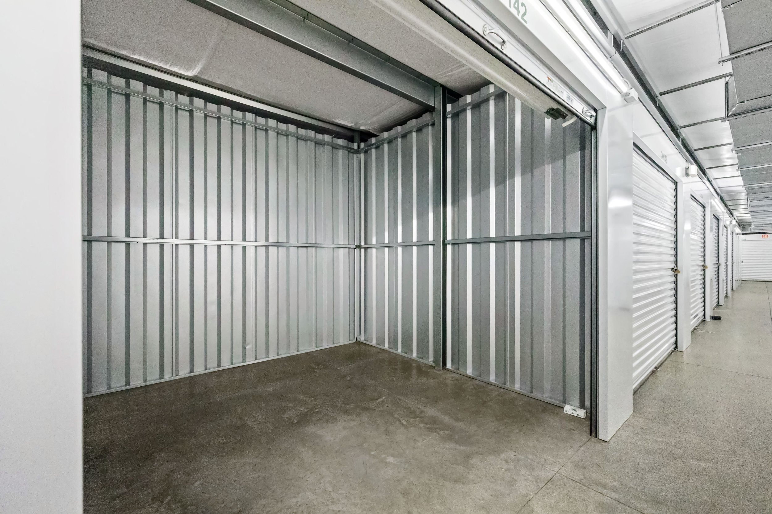 Empty storage unit with metallic walls and a concrete floor in a storage facility, with several other units visible along the hallway.