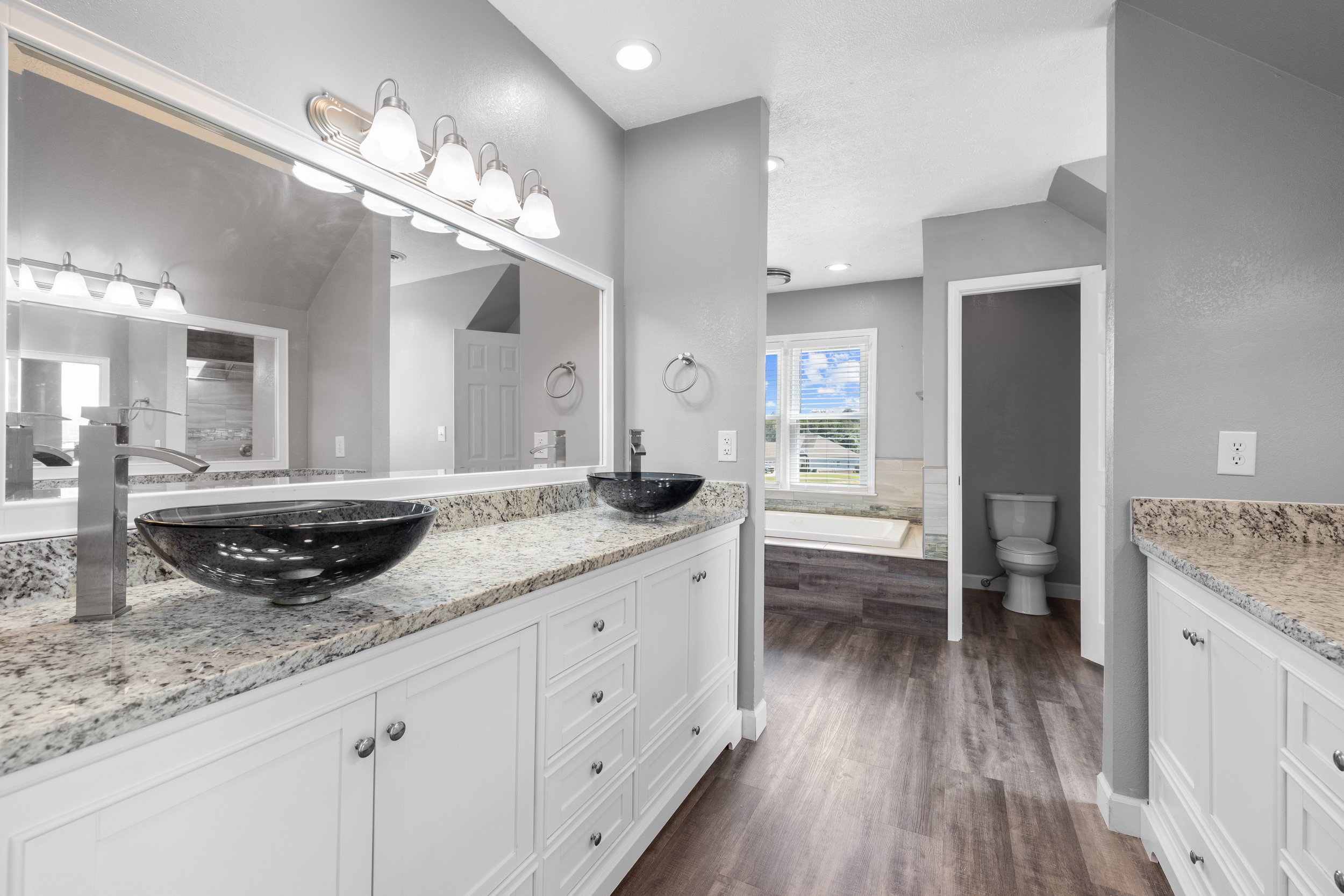 Modern bathroom with double granite countertop vanity, two black vessel sinks, large mirror, wood flooring, toilet in separate room, and bathtub near window.
