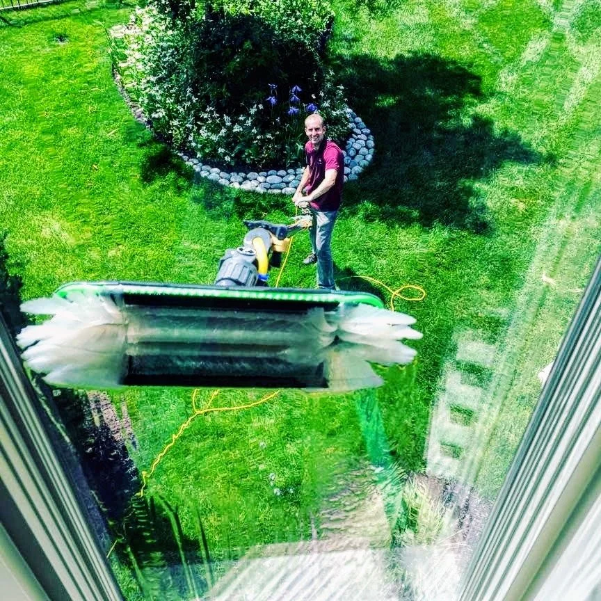 A man wearing a maroon shirt and jeans is standing on a lawn, holding a water fed pole, and cleaning a window in Omaha, Nebraska.