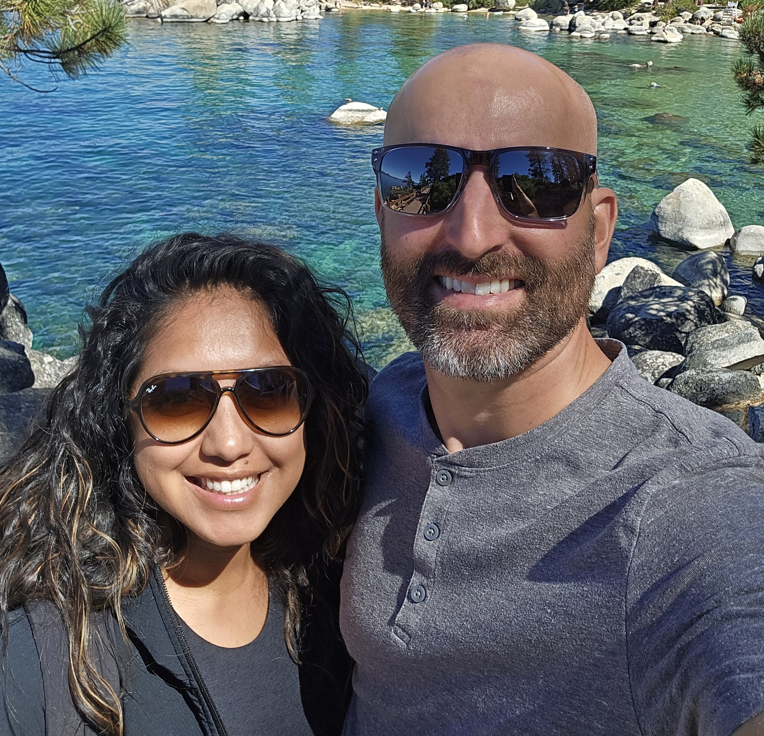 A smiling husband and wife team taking a selfie by a clear blue lake in Omaha, Nebraska.