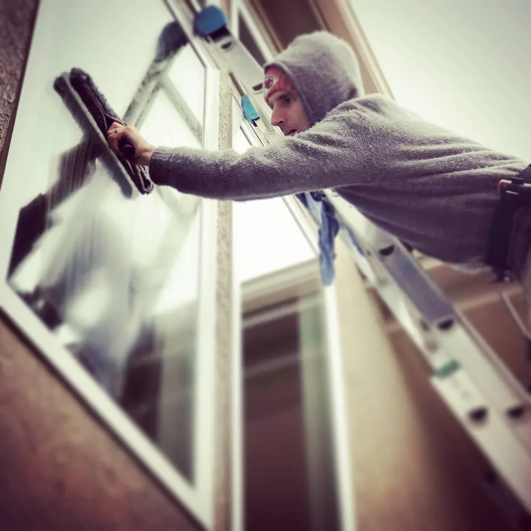 A man cleaning a window in Omaha, Nebraska using a squeegee, wearing a gray hoodie and a gray beanie.