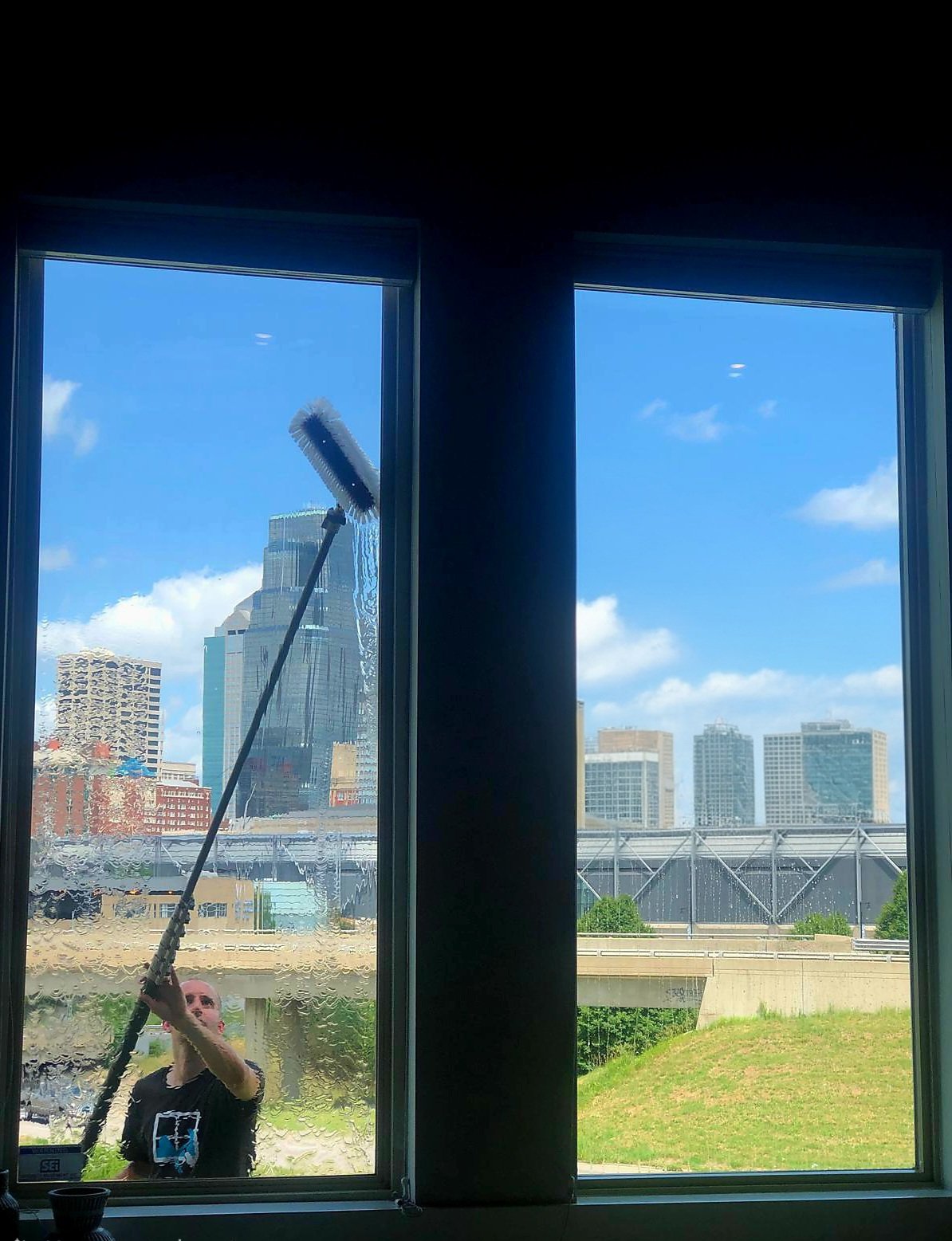 Person cleaning a large window in Omaha, Nebraska on the outside, with city buildings and blue sky visible.